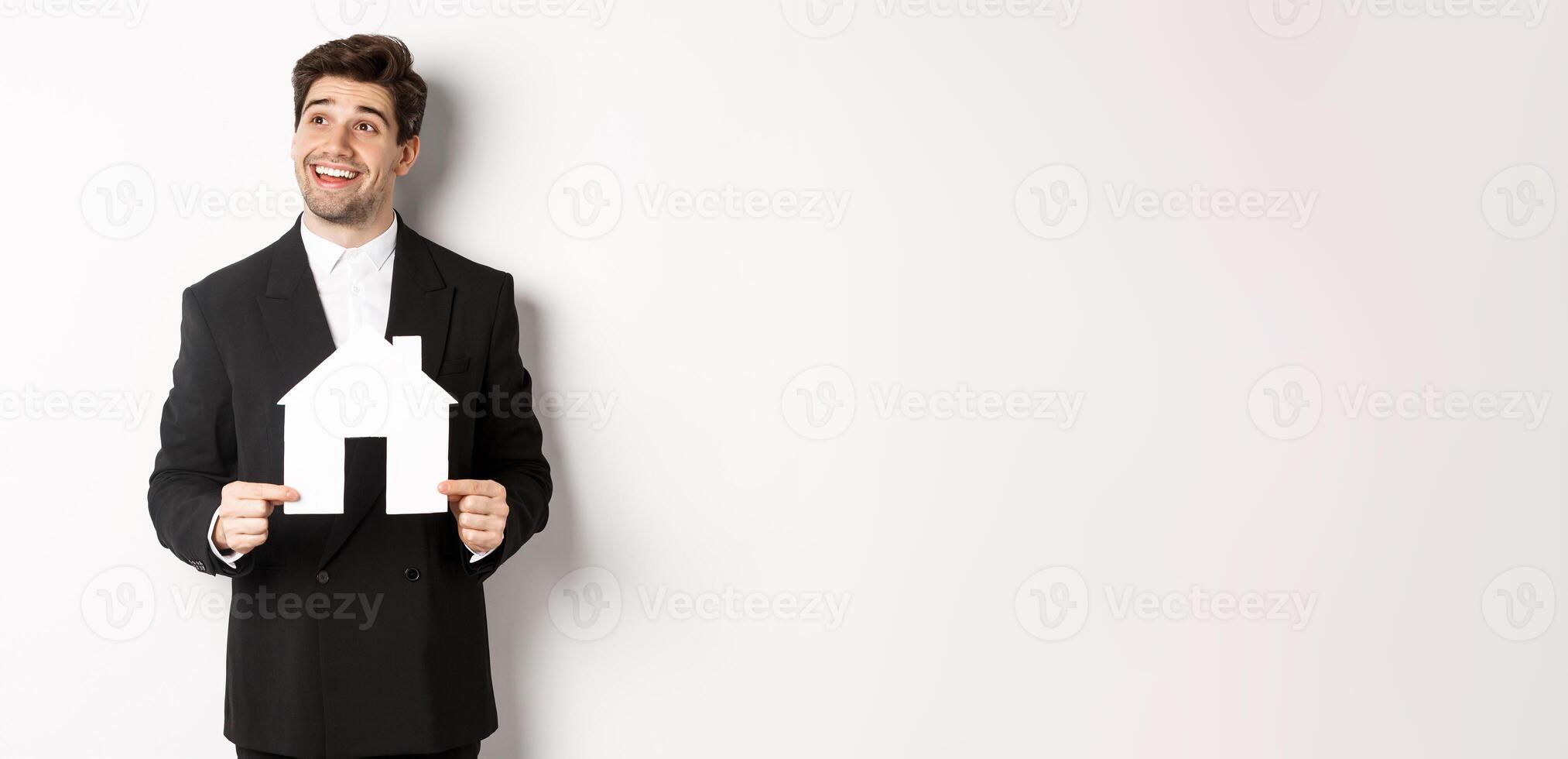 Image of handsome investor in black suit, holding house maket and looking at upper left corner thoughtful, standing against white background photo