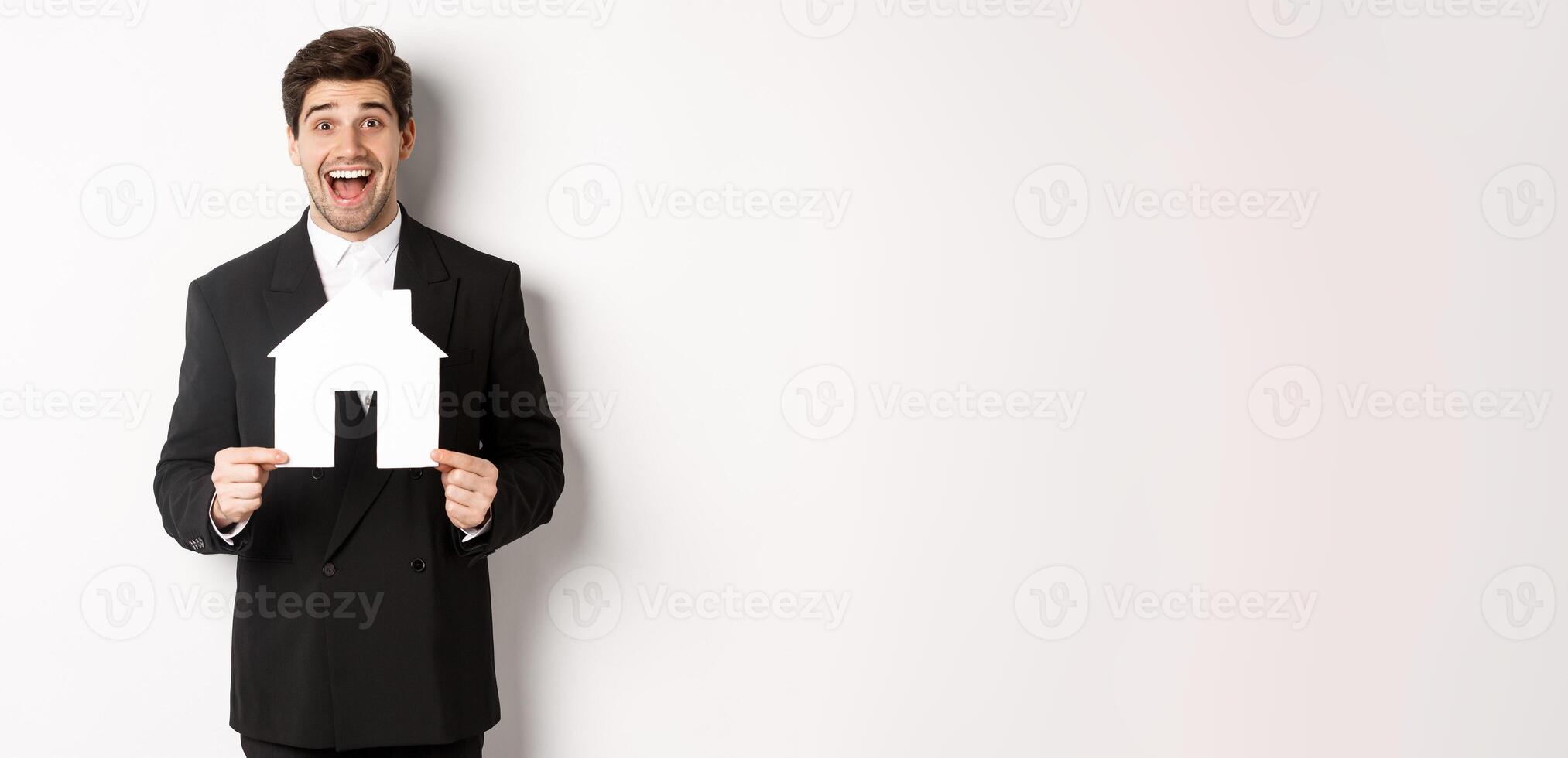 Image of handsome real estate agent in black suit showing home maket, and looking amazed, selling houses, standing against white background photo