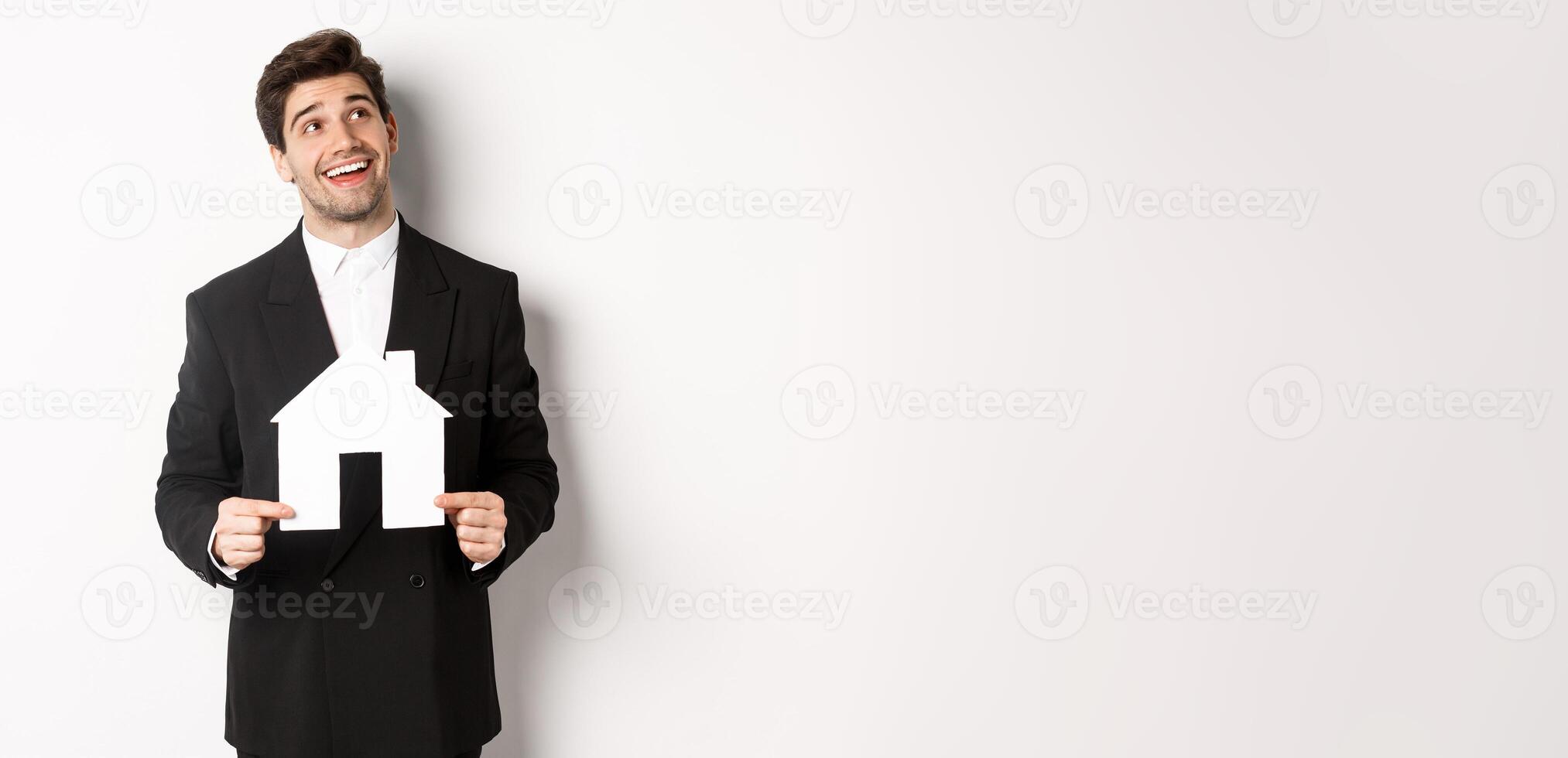 Portrait of handsome man in suit searching for home, holding paper house and looking at upper right corner dreamy, standing over white background photo