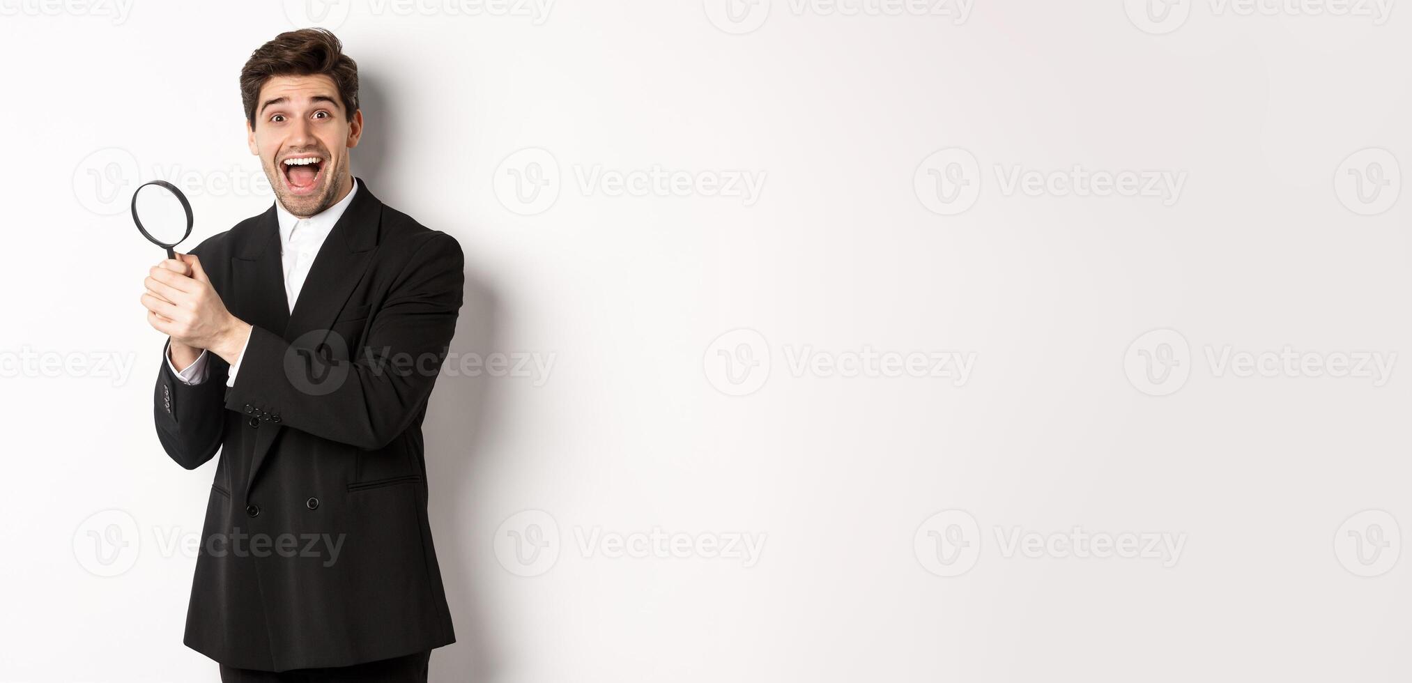 Handsome businessman in black suit, holding magnifying glass and smiling, found something, standing against white background photo