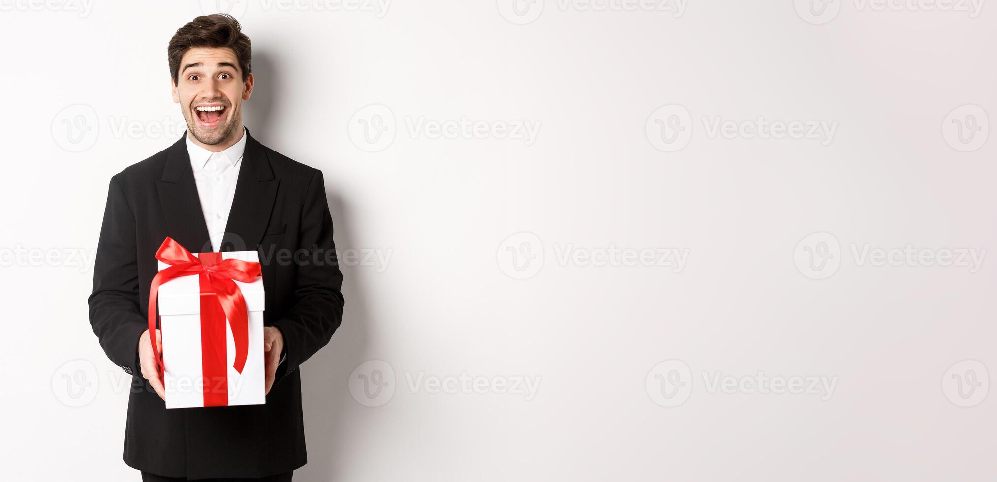 Concept of christmas holidays, celebration and lifestyle. Image of handsome guy in black suit looking excited, have a gift, standing against white background photo