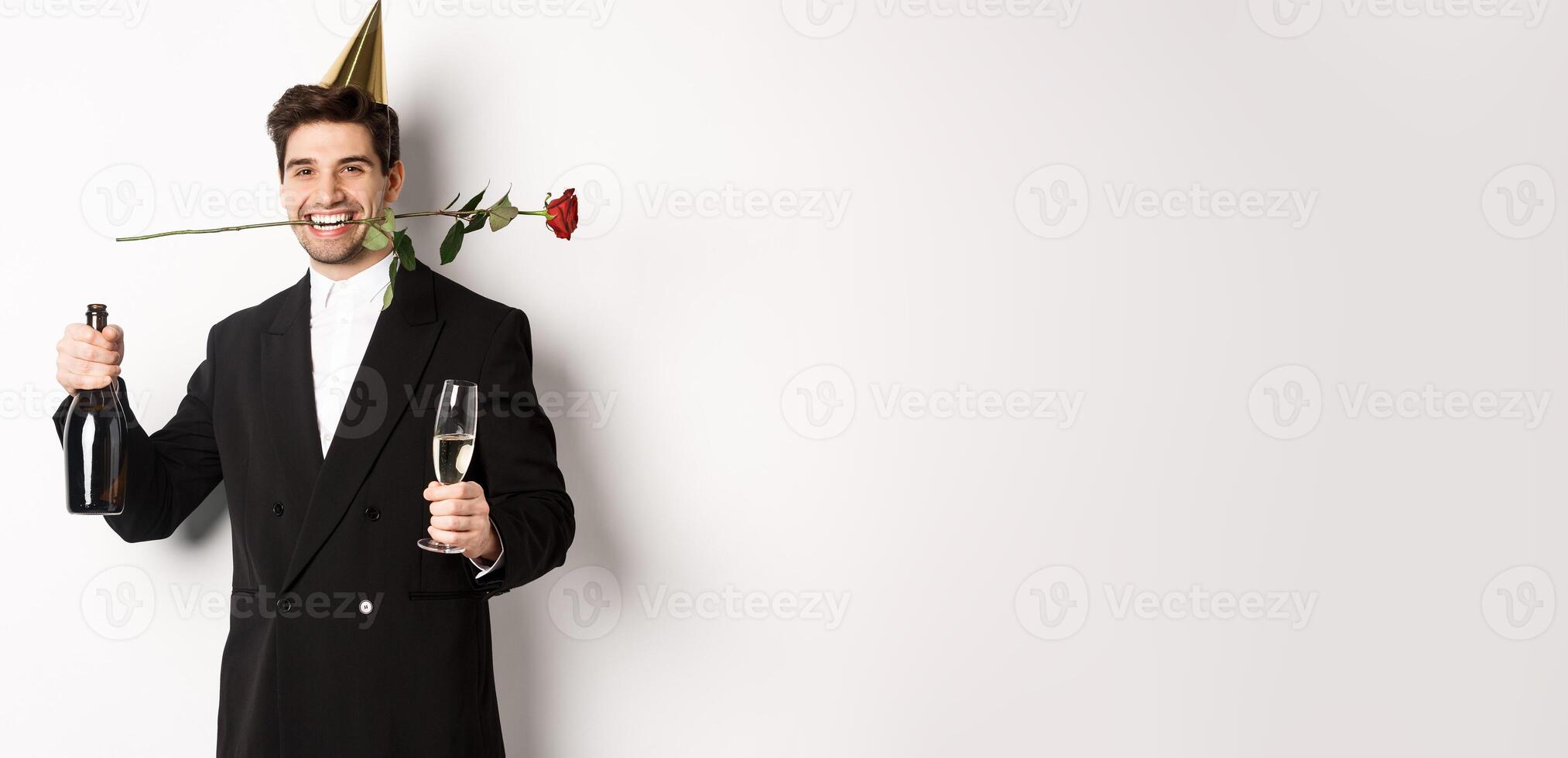Funny guy in trendy suit, celebrating and having a party, holding rose in teeth and champagne, standing over white background photo