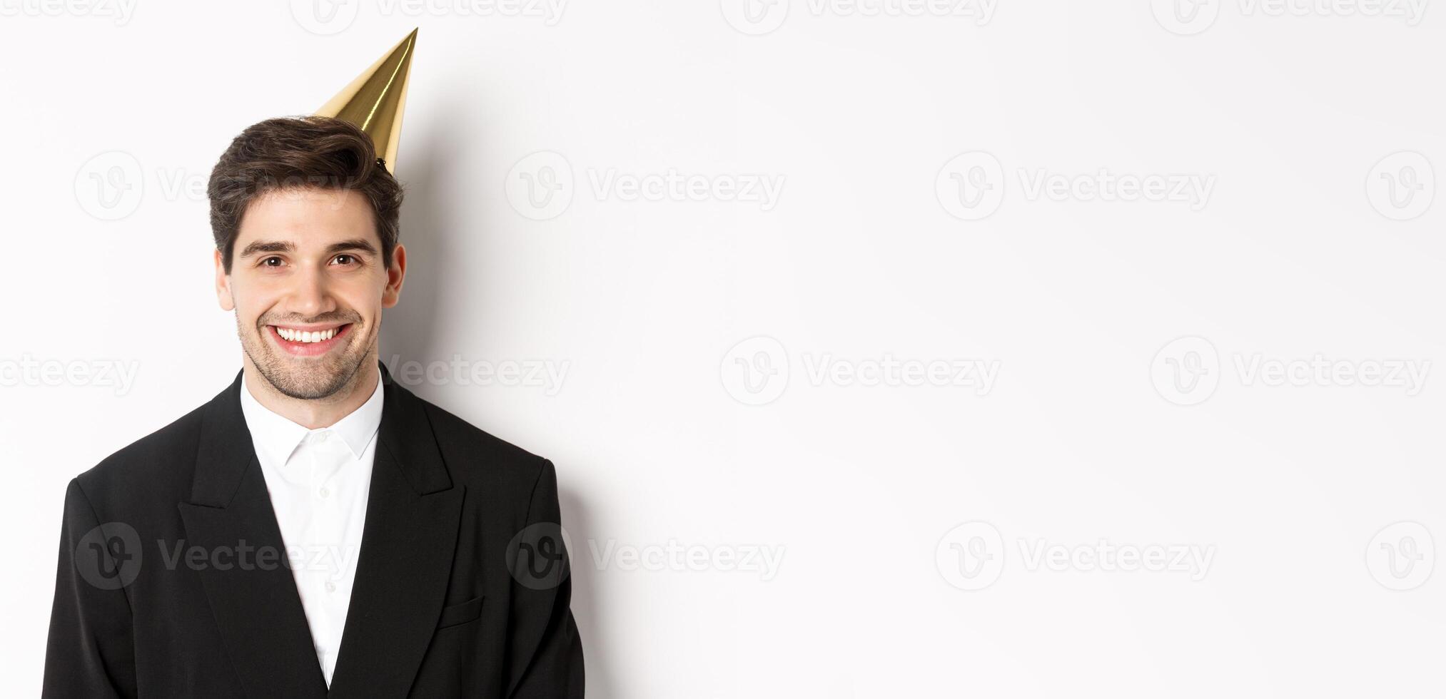 Close-up of handsome happy man in suit and party hat, smiling joyful, celebrating holiday, standing against white background photo