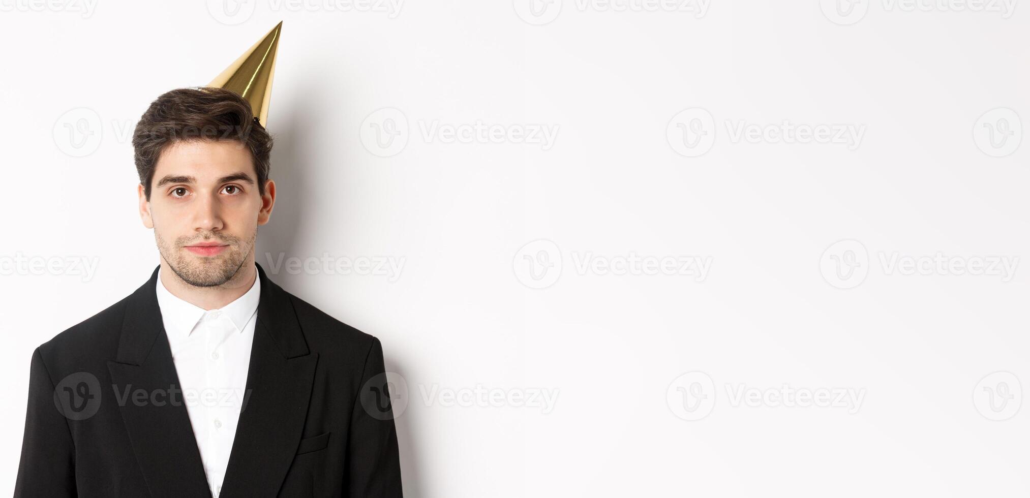 Close-up of handsome man in party hat and trendy suit, celebrating new year, standing against white background photo