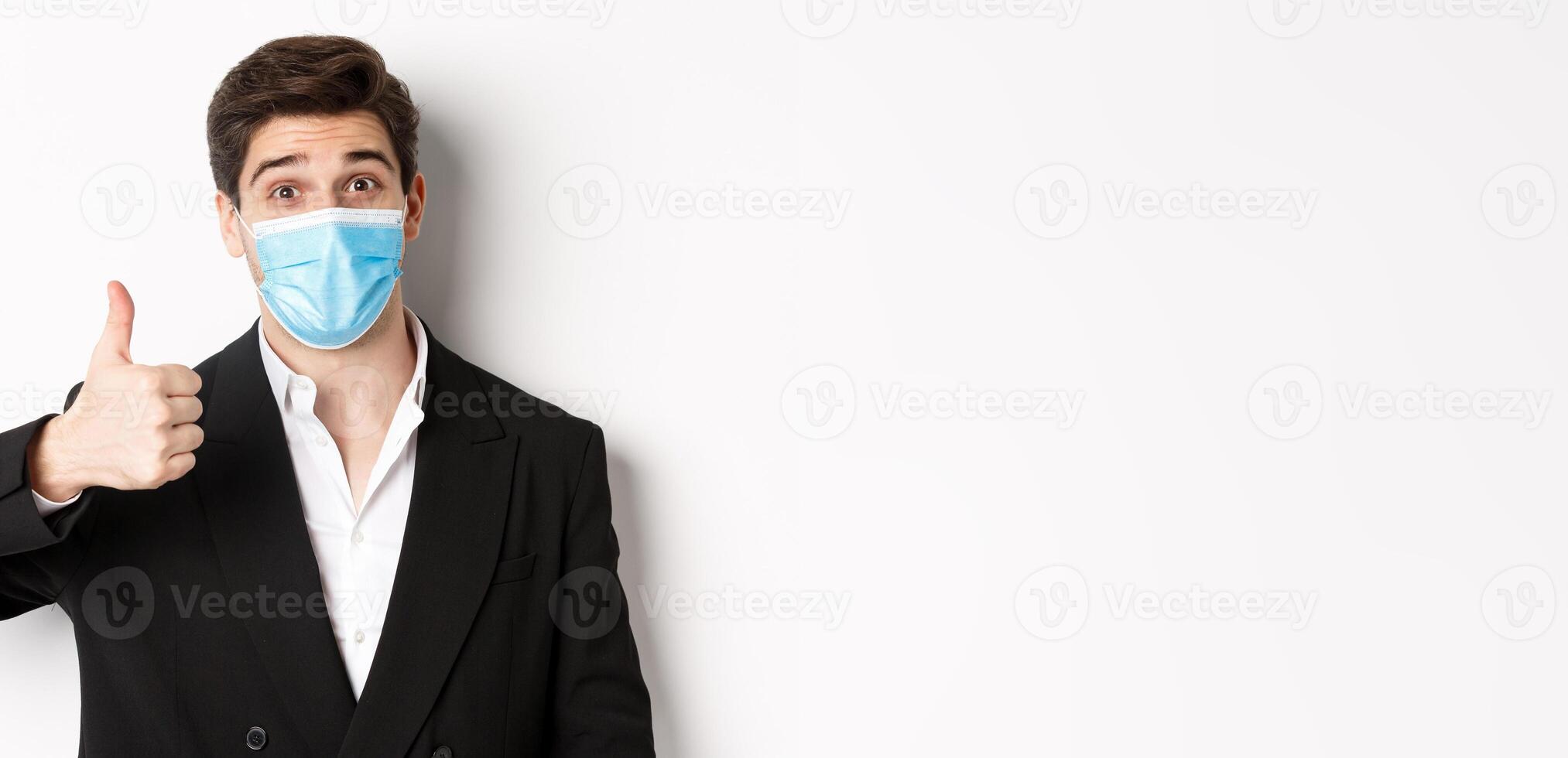 Concept of covid-19, business and social distancing. Close-up of happy businessman in black suit and medical mask, showing thumbs-up, making a compliment, white background photo