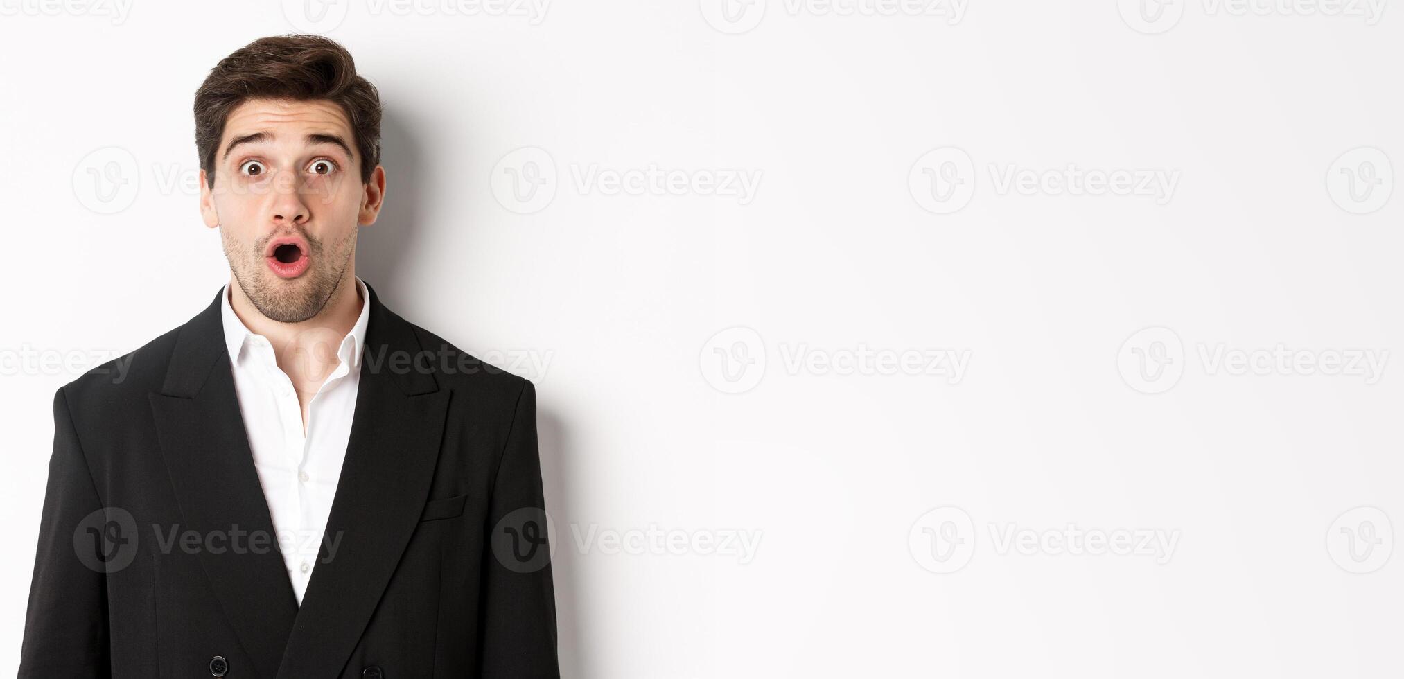 Close-up of attractive man in black suit, looking surprised and impressed at advertisement, standing over white background photo