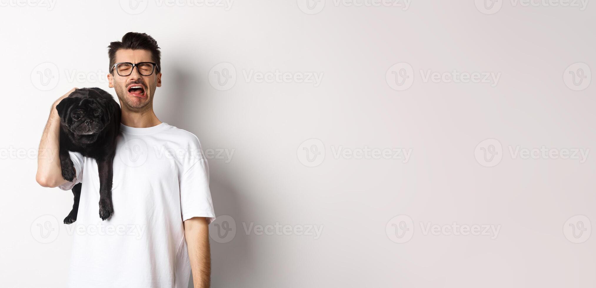Sad dog owner crying, holding cute black pug on shoulder and looking miserable, sobbing while standing over white background photo