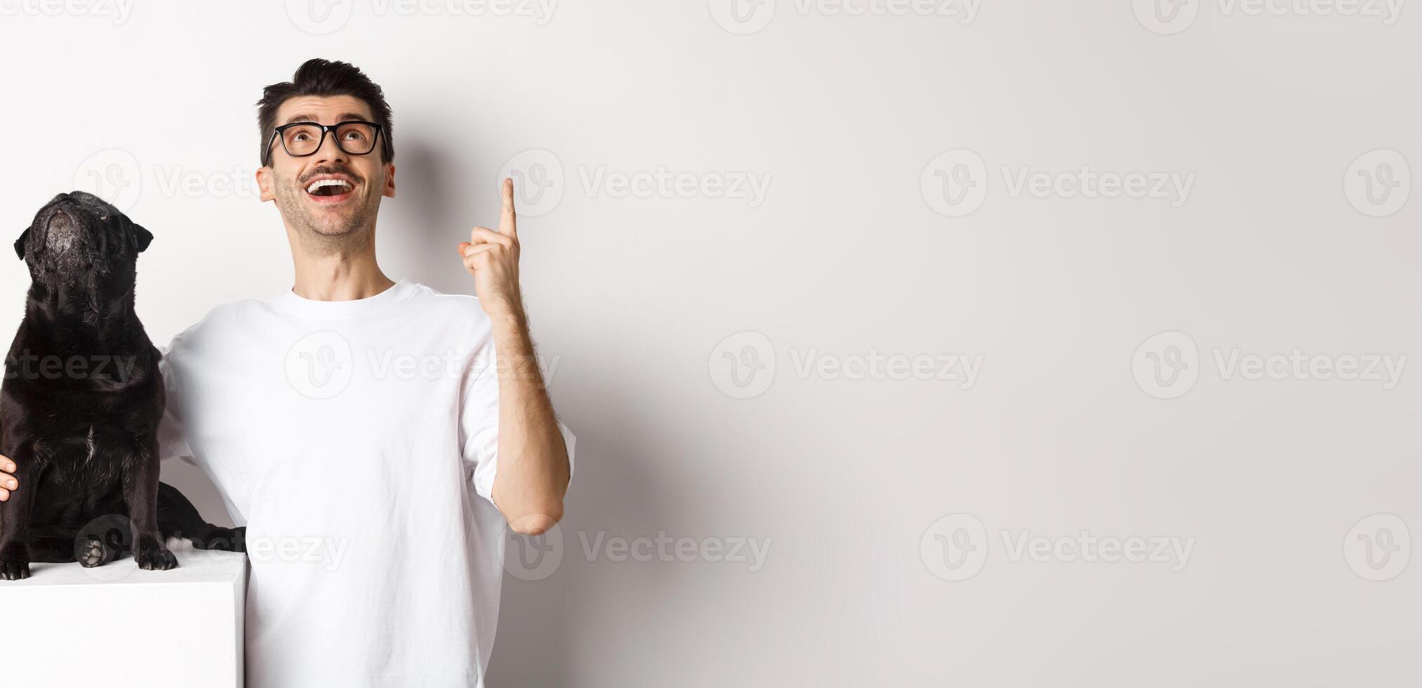 Image of happy young man and dog both looking up at promo, owner pointing finger at top, pug staring at loog, standing over white background photo