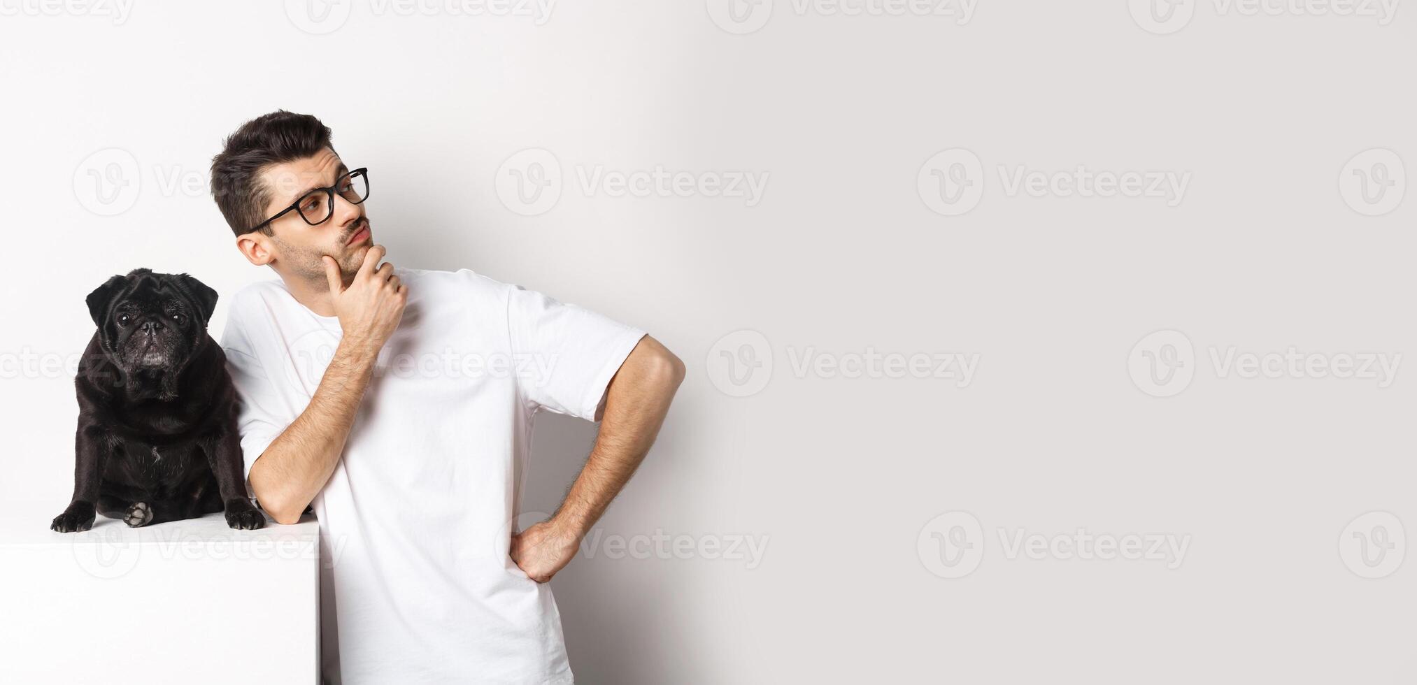 Image of handsome young man, pet owner standing with his dog and looking right at logo, posing with pug against white background photo