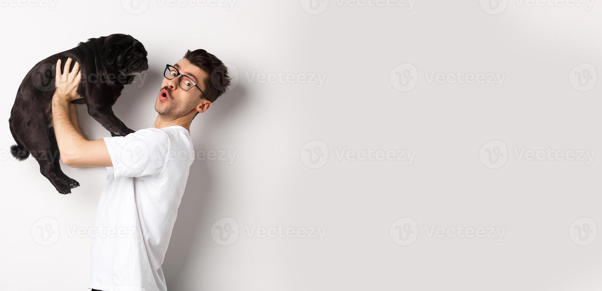 Image of handsome young man loving his pug. Dog owner holding puppy and smiling happy at camera, standing over white background photo
