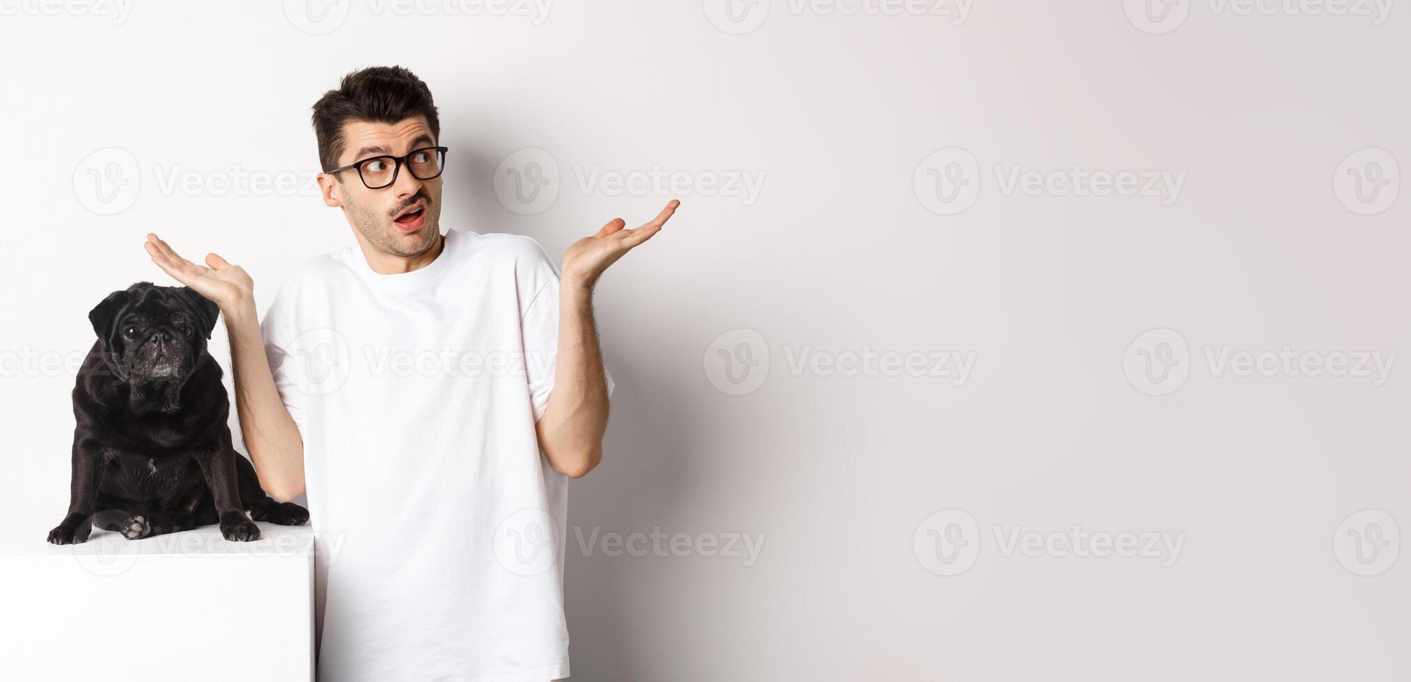 Portrait of confused hipster guy dog owner shrugging, standing near cute black pug pet, white background photo