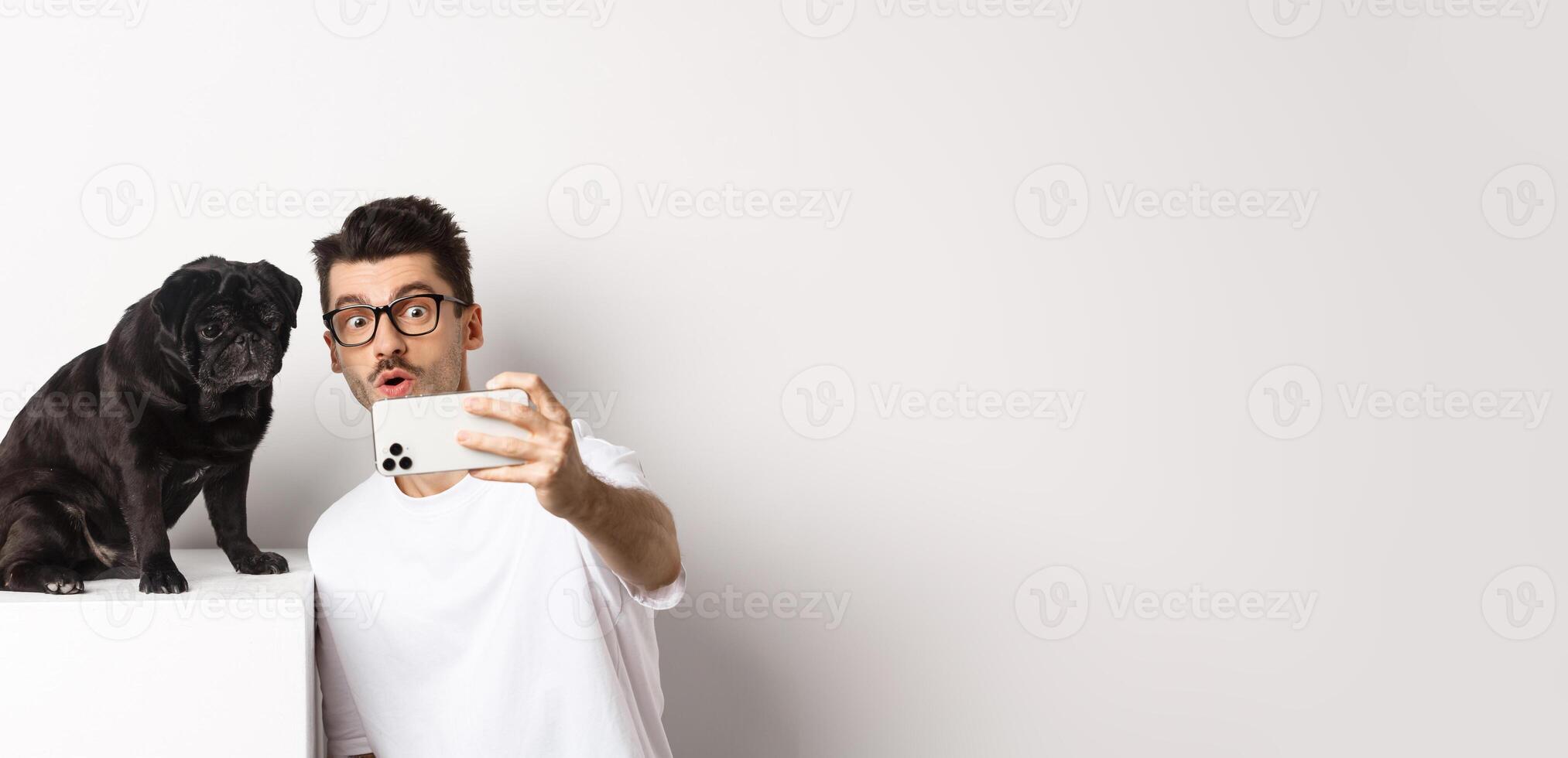 Image of handsome young man taking selfie with cute black dog on smartphone, posing with pug over white background photo