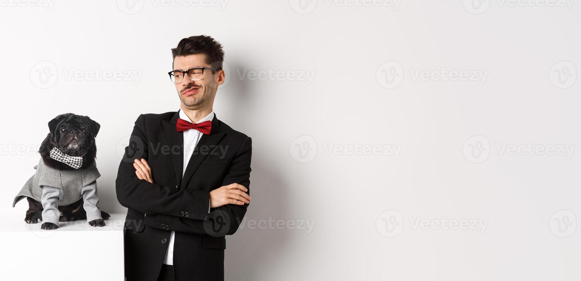 Animals, party and celebration concept. Image of funny young man in suit and glasses, looking skeptical at cute pug in costume, standing over white background photo