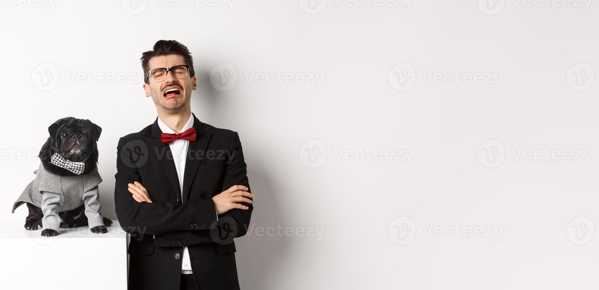 Animals, party and celebration concept. Sad dog owner crying, wearing suit, standing near cute black pug in costume, standing over white background photo