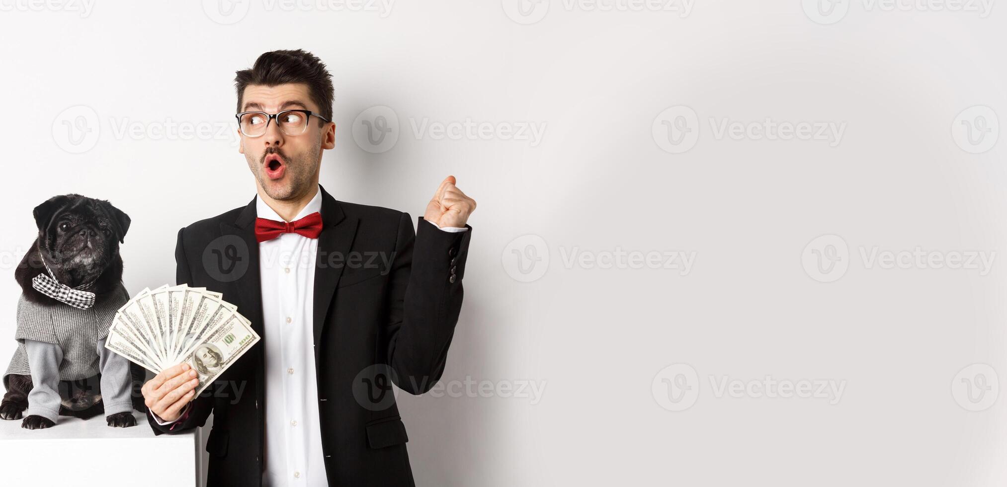 Happy young man in suit earn money with his dog. Guy rejoicing, holding dollars and staring left, black pug in costume staring at camera, white background photo