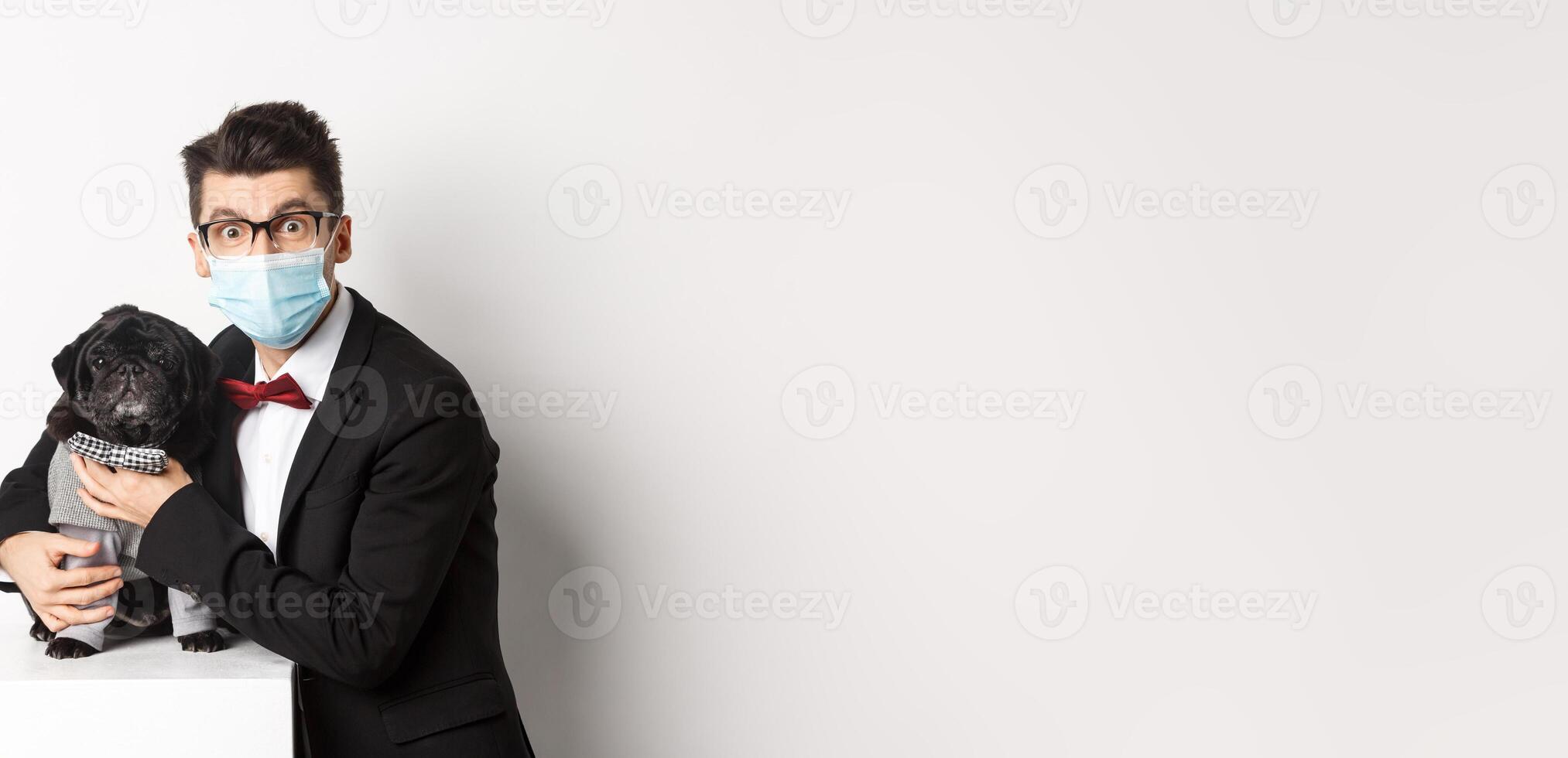 Coronavirus, pets and celebration concept. Happy dog owner in suit and face mask hugging cute black pug in costume, standing over white background photo