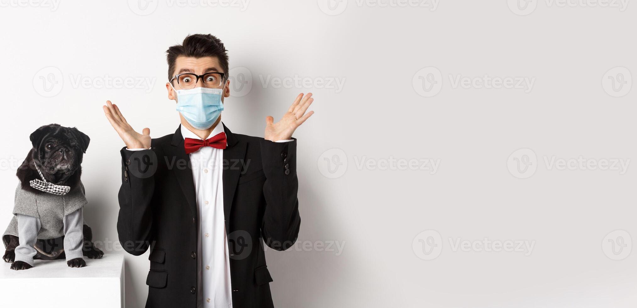 Coronavirus, pets and celebration concept. Amazed young man in face mask and suit staring at camera surprised, cute black dog sitting near owner in party outfit, white background photo
