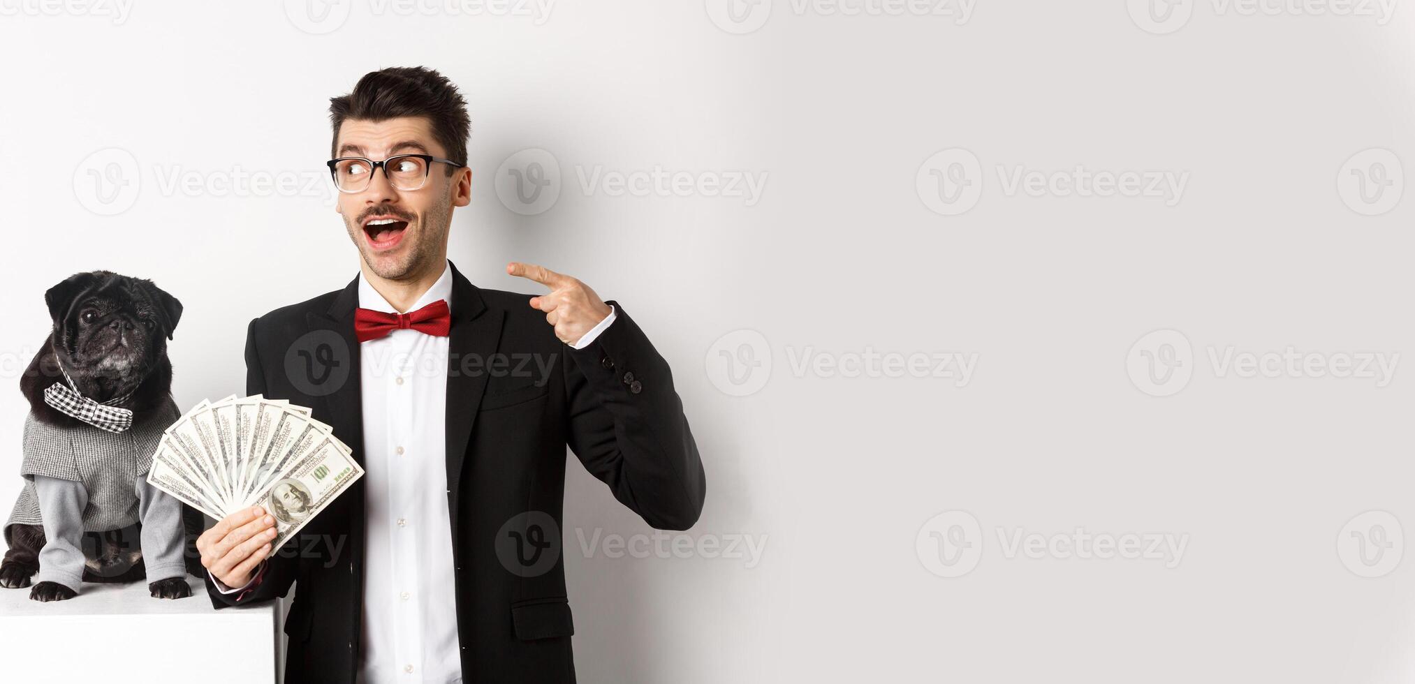 Happy young man in suit earn money with his dog. Guy rejoicing, holding dollars and pointing left, black pug in costume staring at camera, white background photo