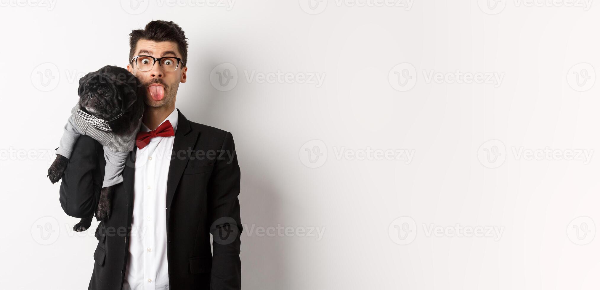 Funny young man in party suit, showing tongue and holding cute black pug on shoulder, celebrating with pet, standing over white background photo