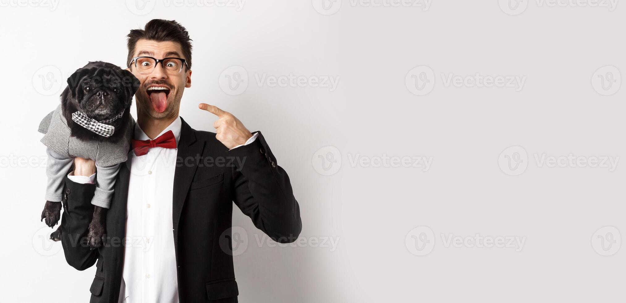 Funny young man in party suit, showing tongue and making grimaces, pointing at cute black dog in winter clothes, standing over white background photo