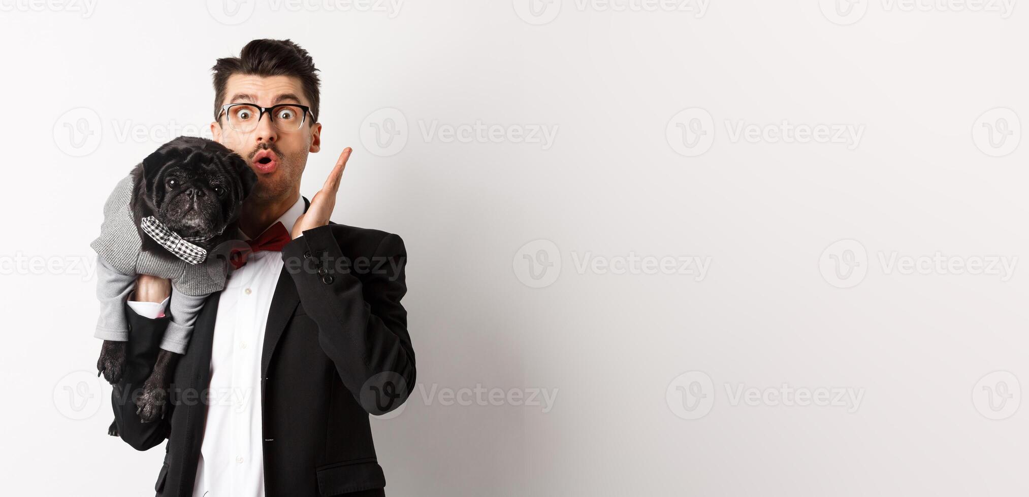 Surprised and amazed dog owner in suit staring at camera, holding cute black pug on shoulder, pet wearing costume, posing over white background photo