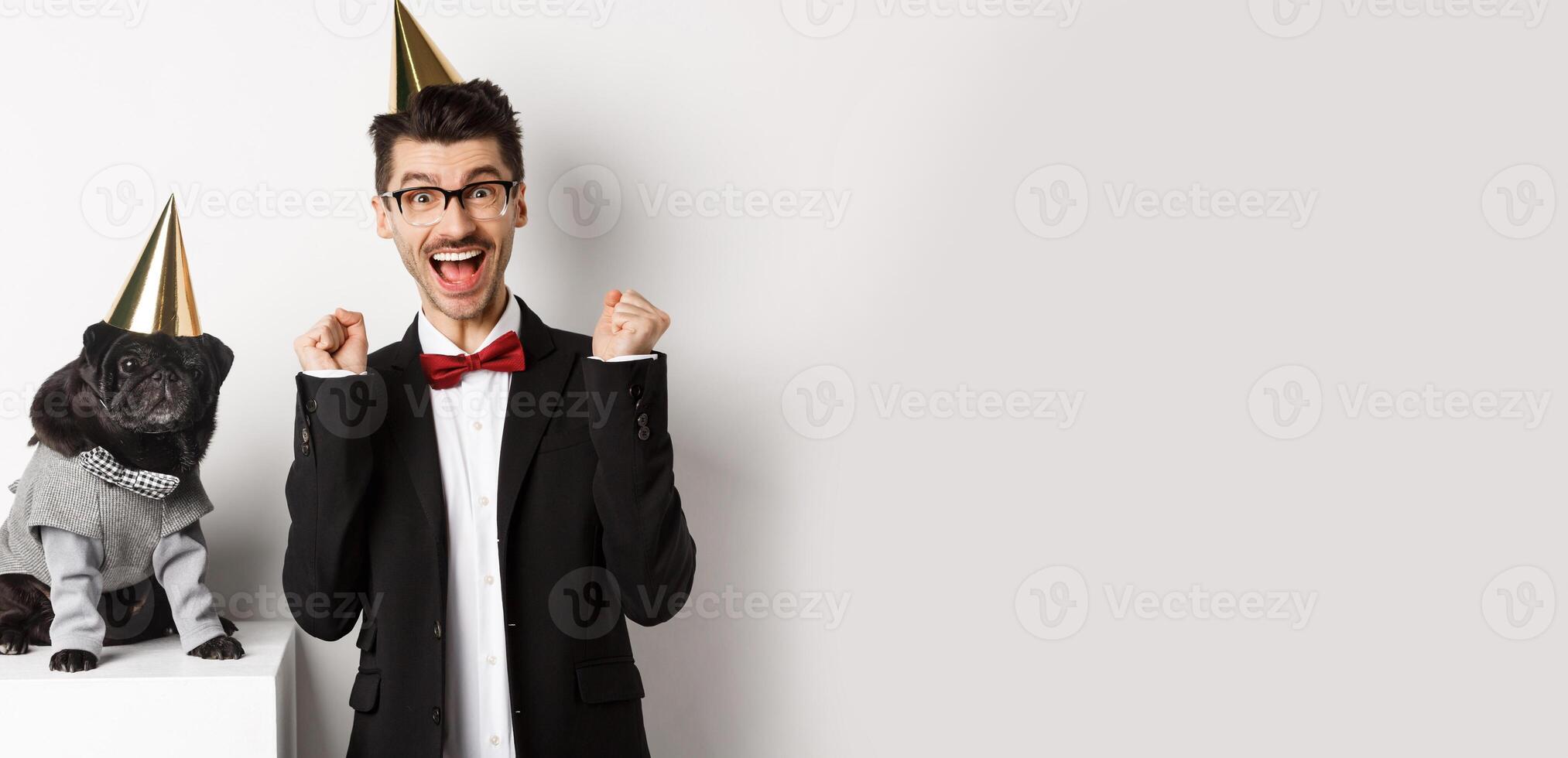 Cheerful young man shouting for joy, dog and owner wearing birthday party cones and celebrating, guy rejoicing and staring at camera, white background photo
