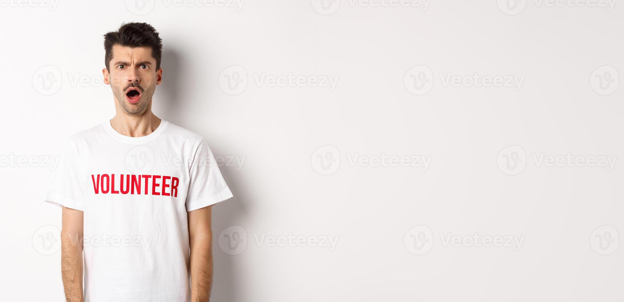 Shocked and confused man in volunteer t-shirt staring at camera and frowning displeased, standing against white background photo