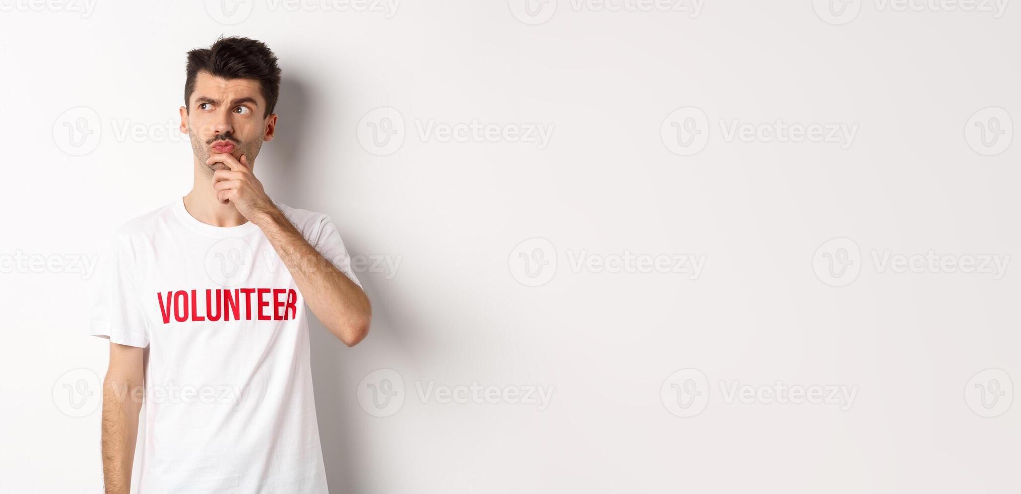 Thoughtful young male volunteer in white t-shirt searching solutions, looking at upper left corner and thinking, standing over studio background photo