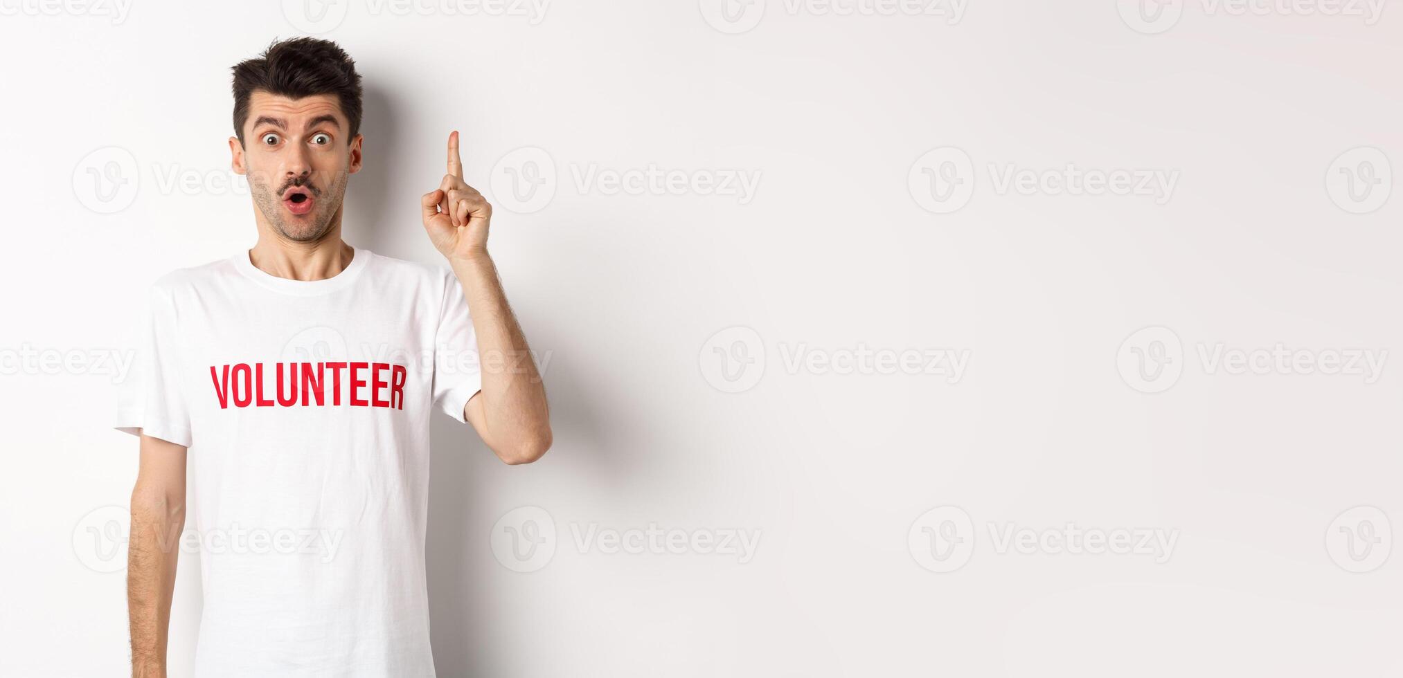 Handsome young man in volunteer t-shirt having an idea, raising finger and saying suggestion, pointing up, standing over white background photo