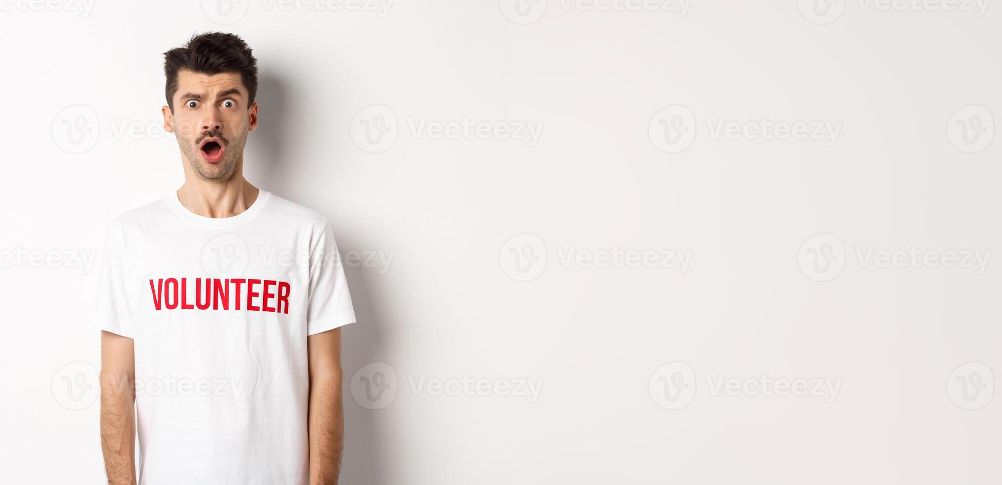 Shocked and confused man in volunteer t-shirt staring at camera speechless, standing against white background photo