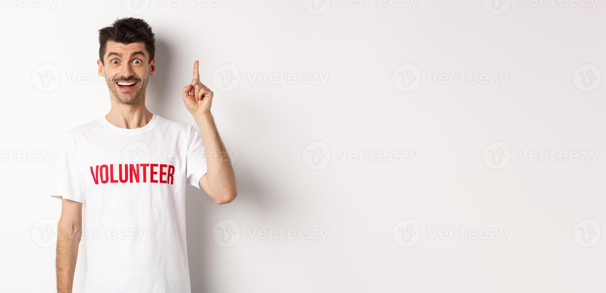 Handsome young man in volunteer t-shirt having an idea, raising finger and saying suggestion, pointing up, standing over white background photo