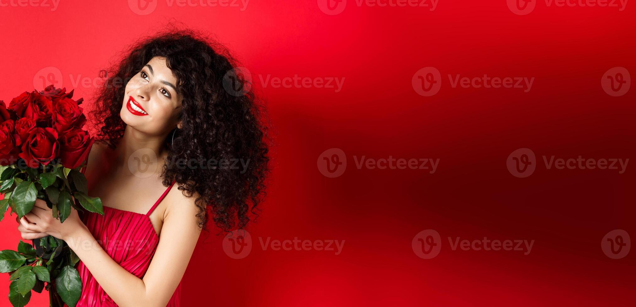 Happy beautiful woman in dress, holding flowers and smiling romantic, looking aside at logo, standing against red background photo