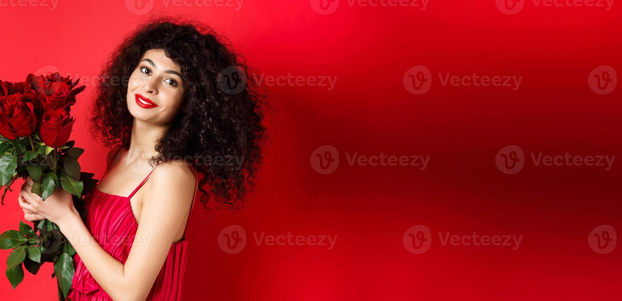 Happy beautiful woman in dress, holding flowers and smiling romantic, standing against red background photo