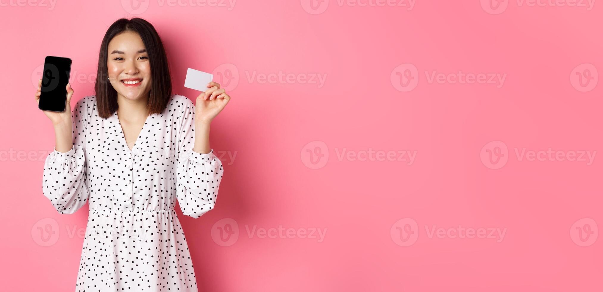 Joyful korean woman showing smartphone screen and credit card, paying for internet order, demonstrating online shopping app, standing over pink background photo