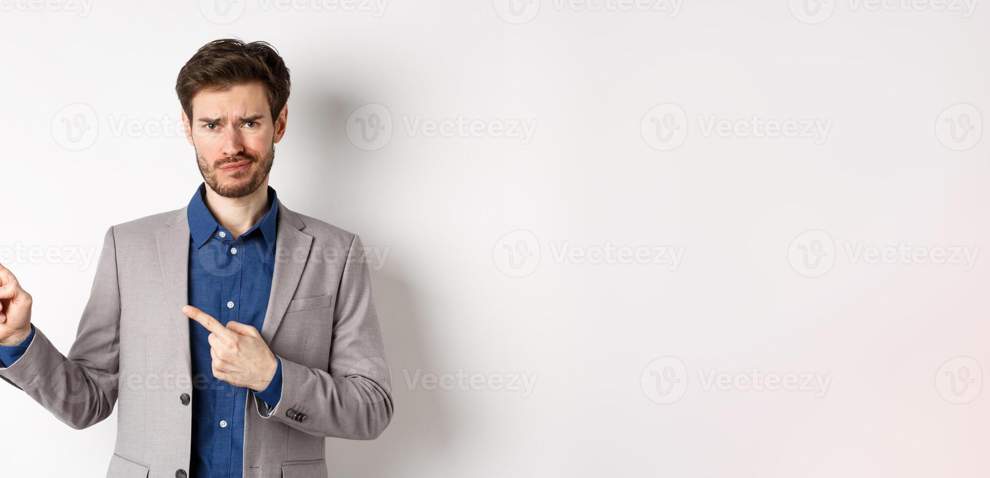 Bad idea. Displeased skeptical guy in business suit smirk and frown, pointing fingers left at upsetting news, looking hesitant at camera, white background photo