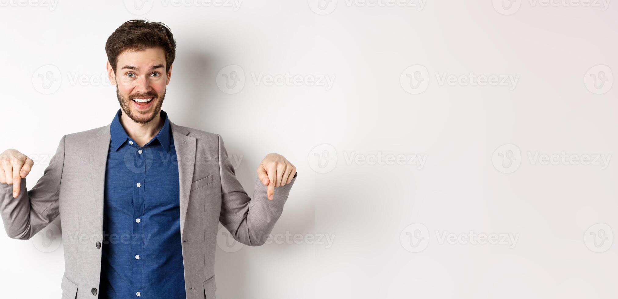 Hey check this out. Happy businessman in suit pointing fingers down and smiling, inviting to event, showing link, standing on white background photo