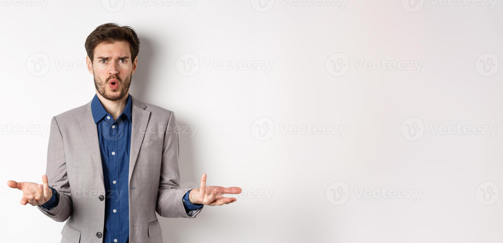 Confused and shocked businessman raising hands up and asking what happened, frowning disappointed, waiting for explanation, standing in suit on white background photo