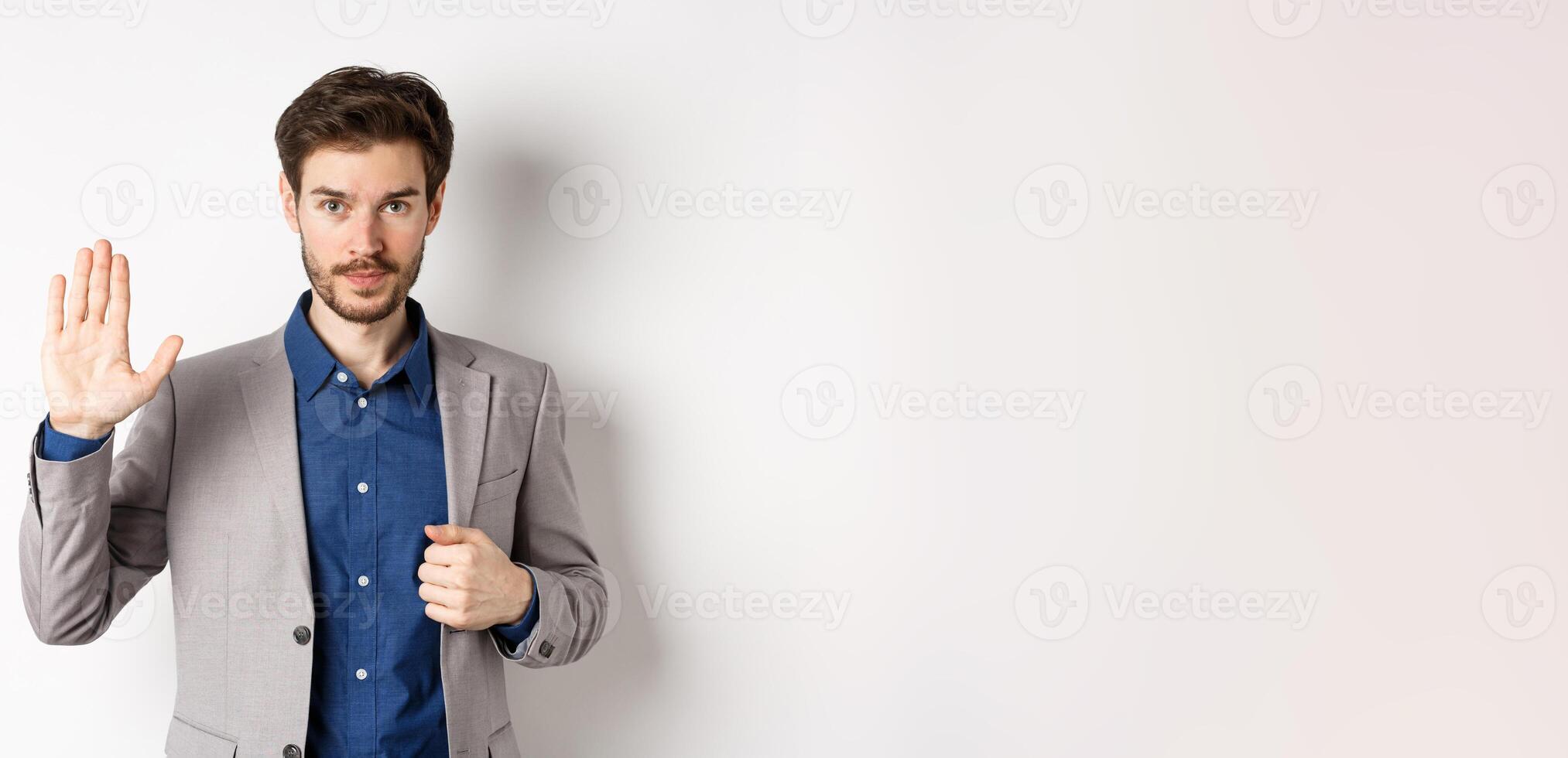 Businessman in formal suit raising hand up to say hello, contactless greeting, smiling friendly, standing on white background photo
