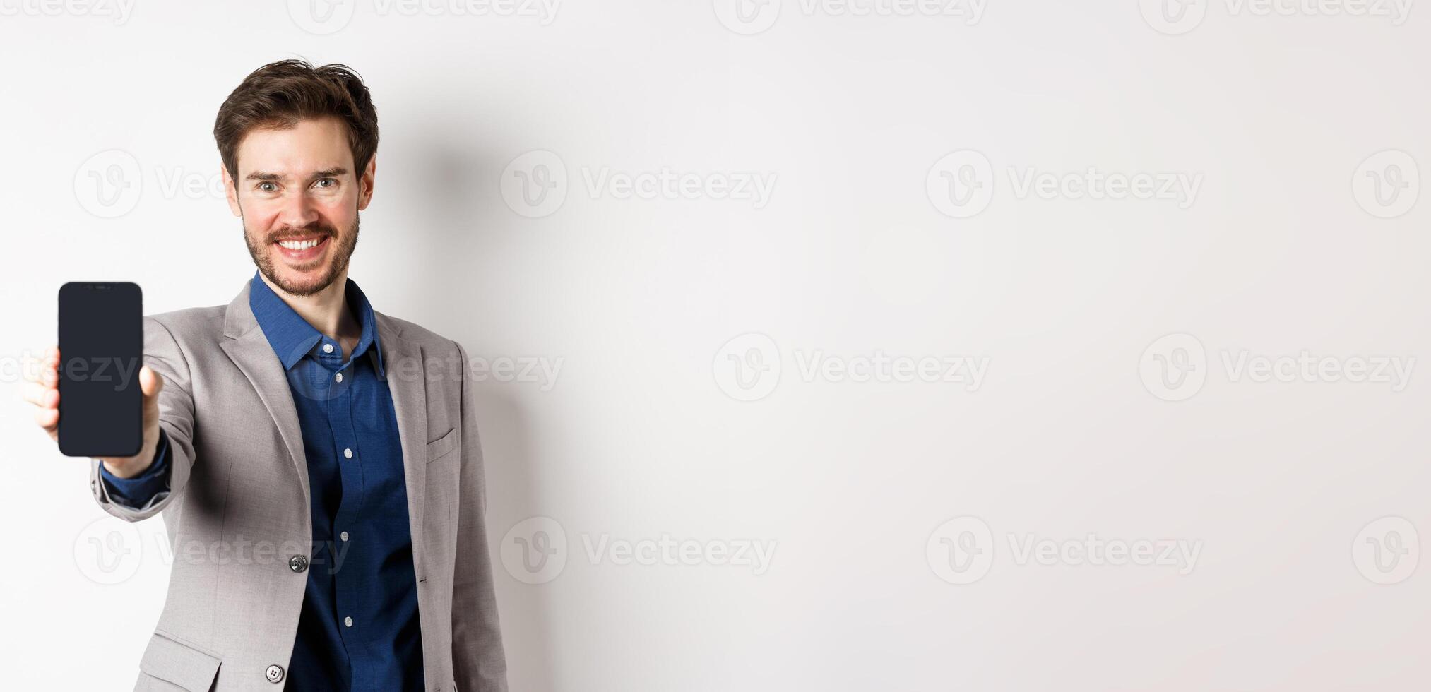 E-commerce and online shopping concept. Confident businessman in suit stretch out hand with empty smartphone screen, showing on phone, standing against white background photo