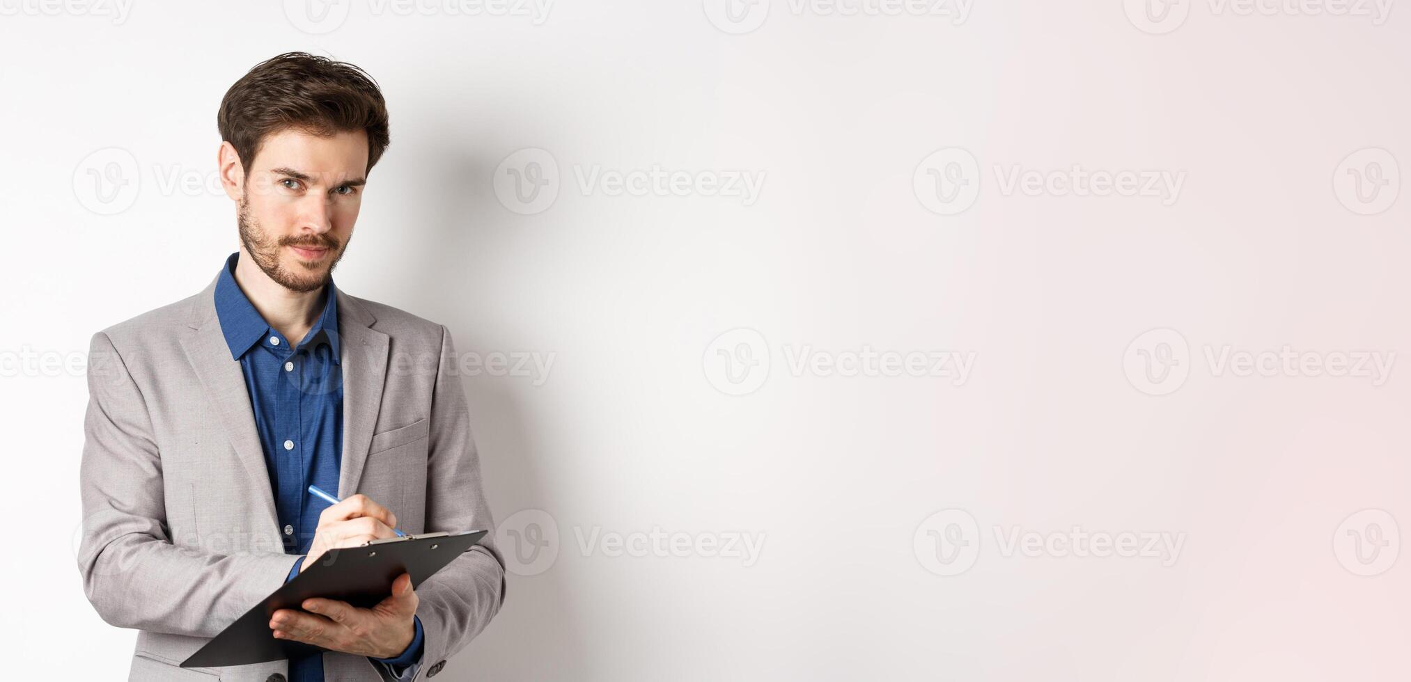 Successful businessman in grey suit taking notes, working in office clothing, holding clipboard and looking at camera, white background photo
