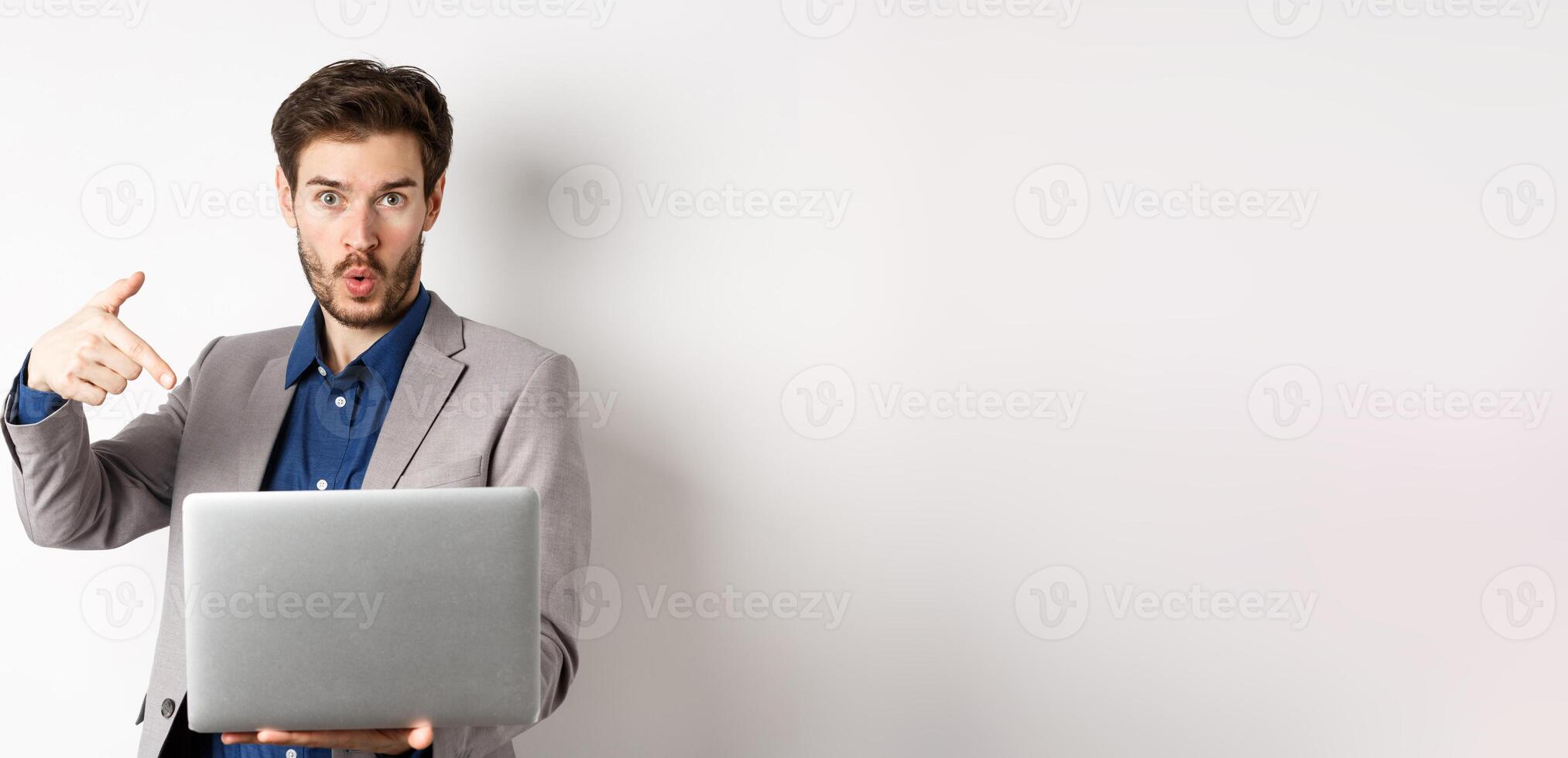Excited handsome man asking to look here, pointing at laptop screen with amazed face, standing in business suit against white background photo