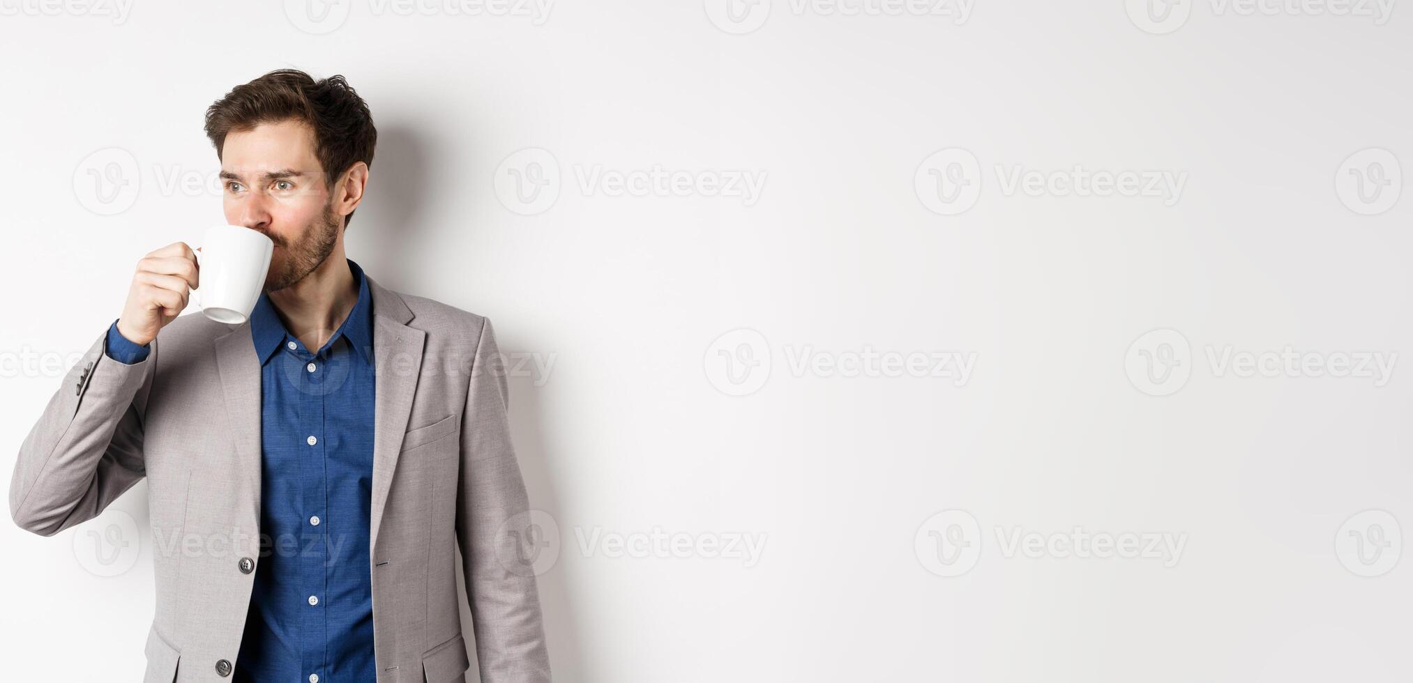 Handsome businessman in suit drinking coffee or tea from office mug, looking aside at logo, standing against white background photo