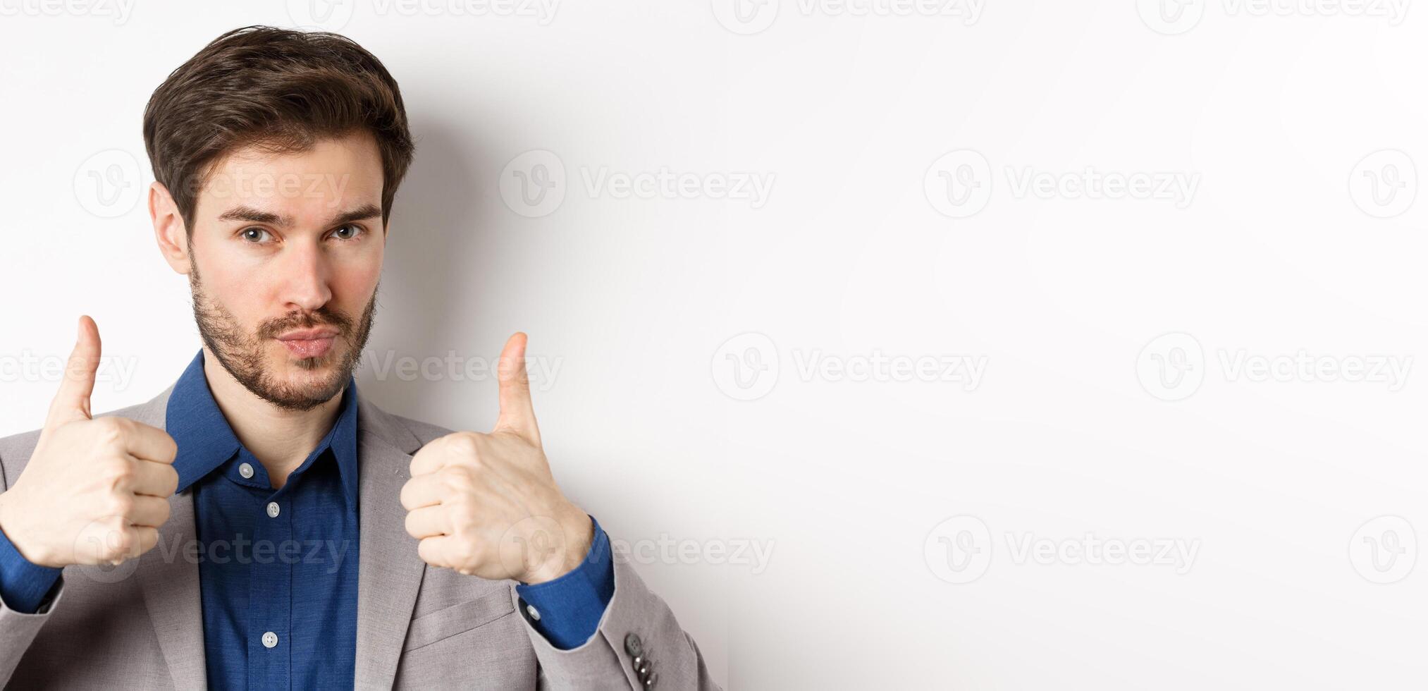Well done. Satisfied businessman in suit showing thumbs up in approval, like and agree, recommend good company, white background photo