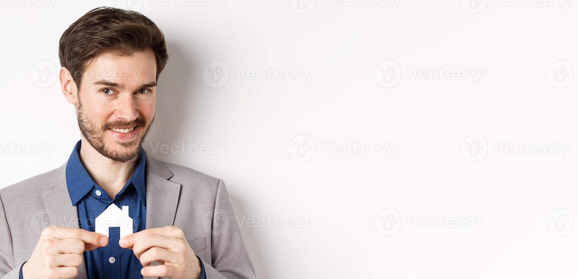 Real estate and insurance concept. Smiling male realtor showing small paper house cutout and looking at camera, selling property, white background photo