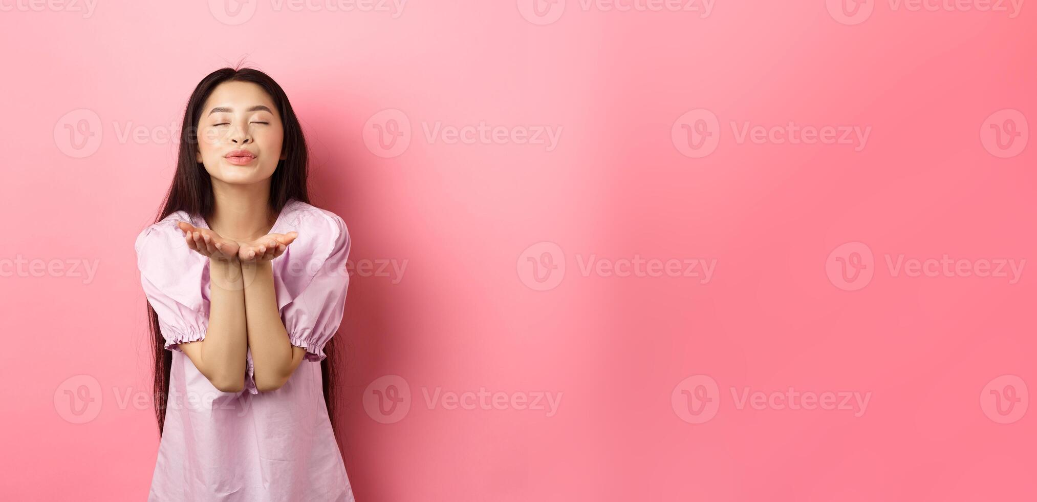 Cute and romantic asian girl blowing air kiss at camera, close eyes and smile tenderly, standing in dress against pink background photo