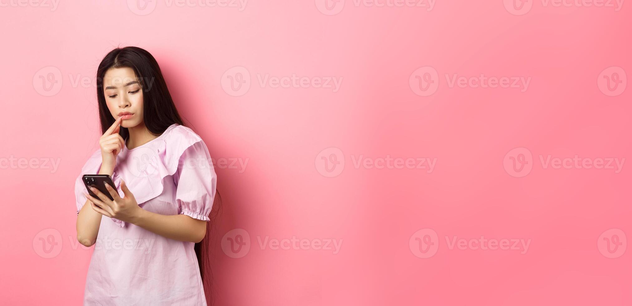 Online shopping. Pensive asian girl looking at smartphone screen, making choice, standing in dress against pink background photo