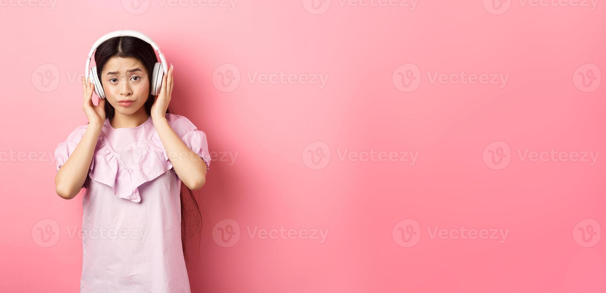 Skeptical girl unamused with song, listening music in headphones and frowning displeased, standing against pink background photo