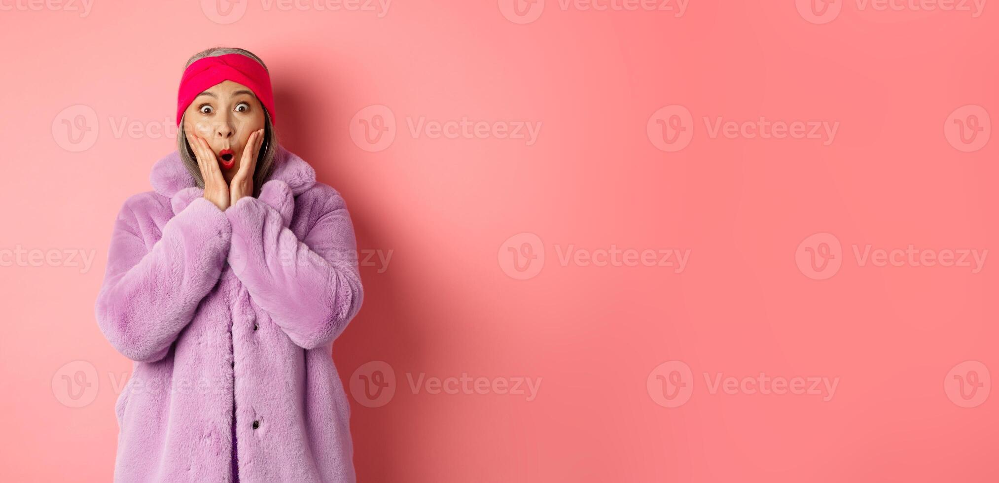 Fashion concept. Trendy old asian woman looking surprised at camera. Mature female in purple coat and headband looking amazed at camera, checking out promotion on pink background photo