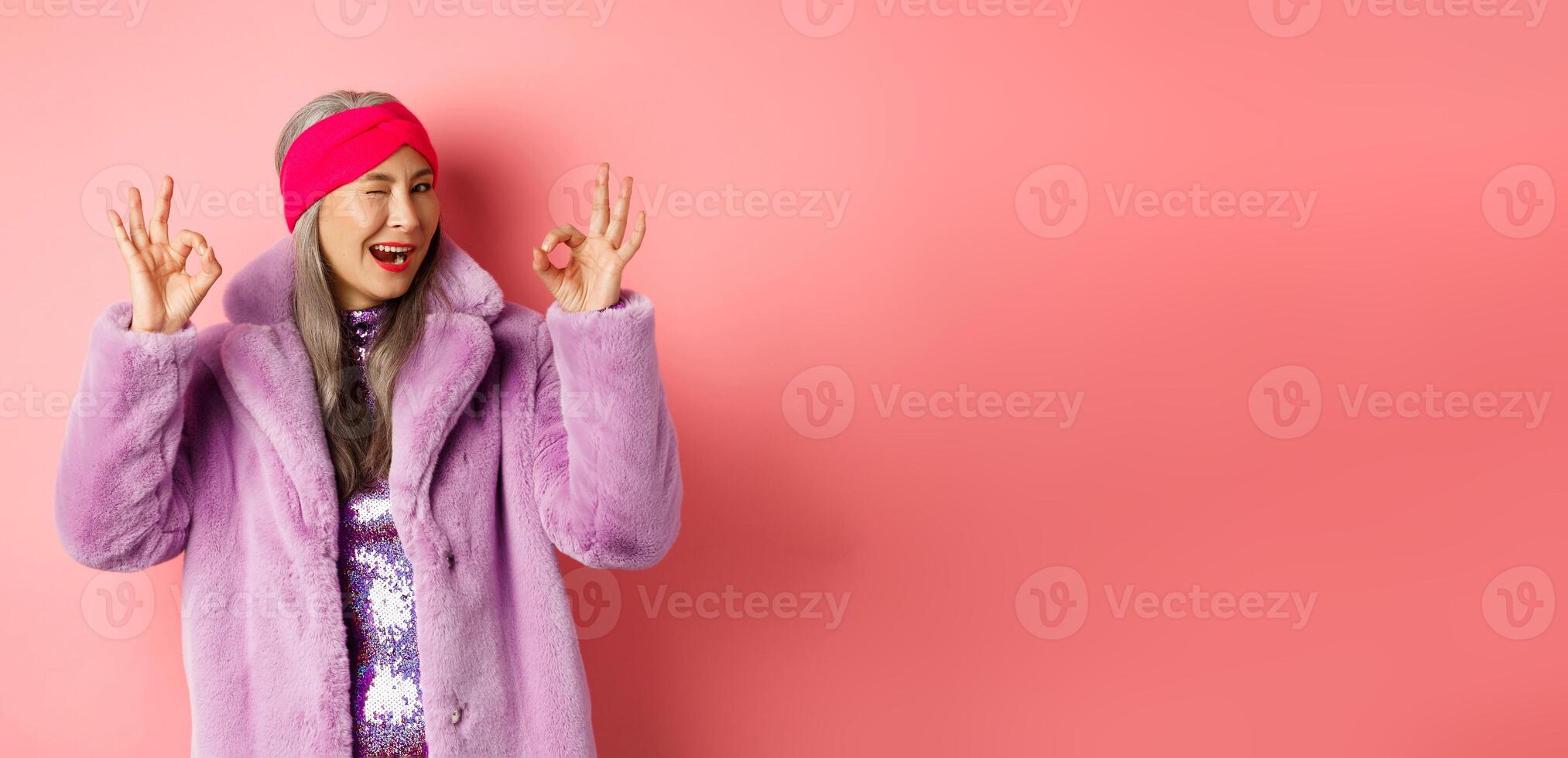 Fashion and shopping concept. Stylish asian granny saying all okay and winking. Trendy senior woman in winter fake-fur coat show OK sign, praising good product, pink background photo