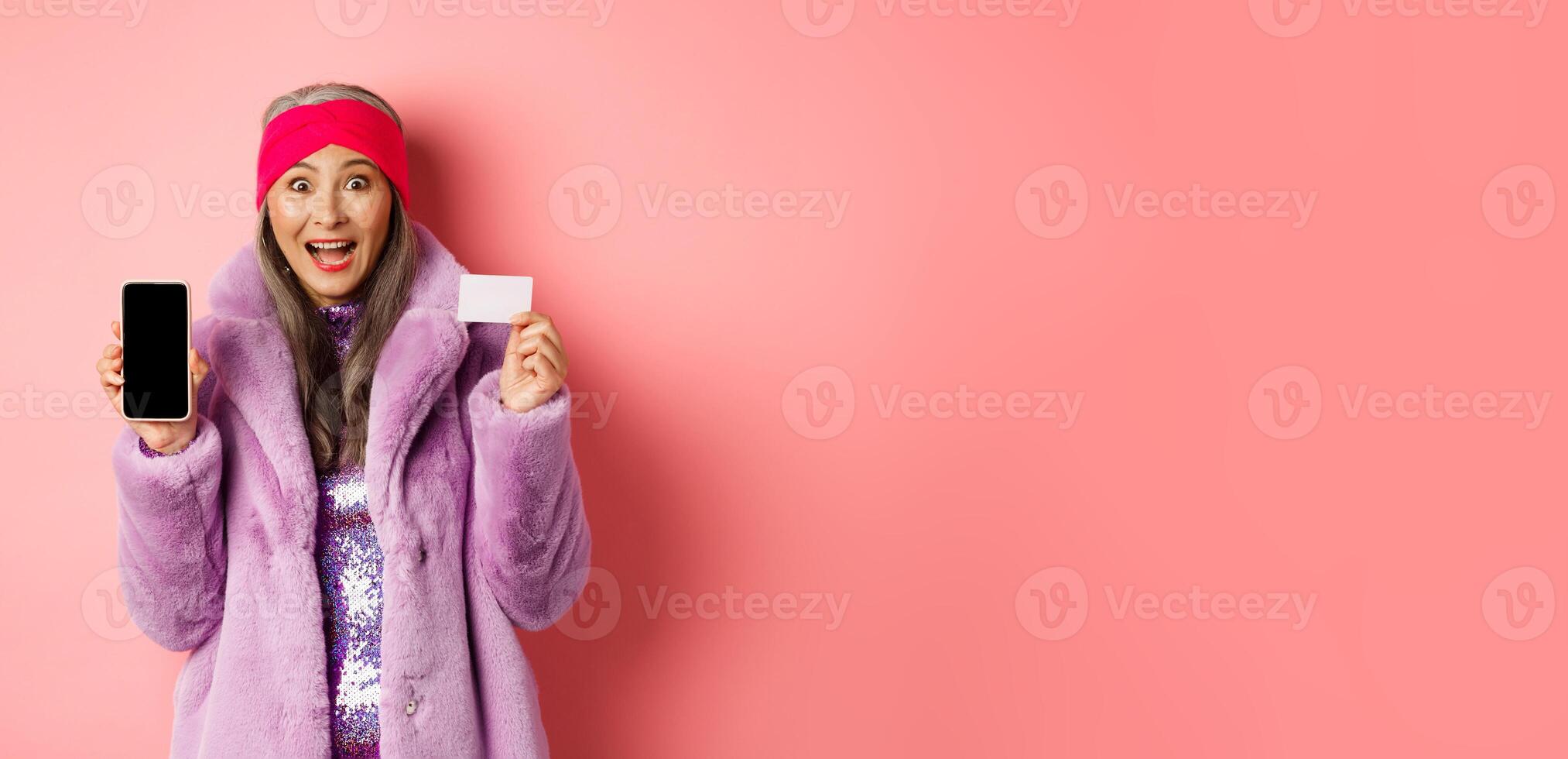 Special promotion. Excited asian senior woman showing blank smartphone screen and plastic credit card, checking out online offer, pink background photo
